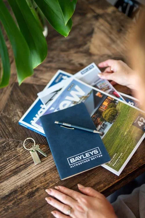 A wooden table with a green plant in the top left corner. On the table are magazines, a blue booklet labeled 'Bayleys' with the slogan 'Altogether Better,' a pen resting on the booklet, a set of keys, and two hands, one holding a magazine and the oth