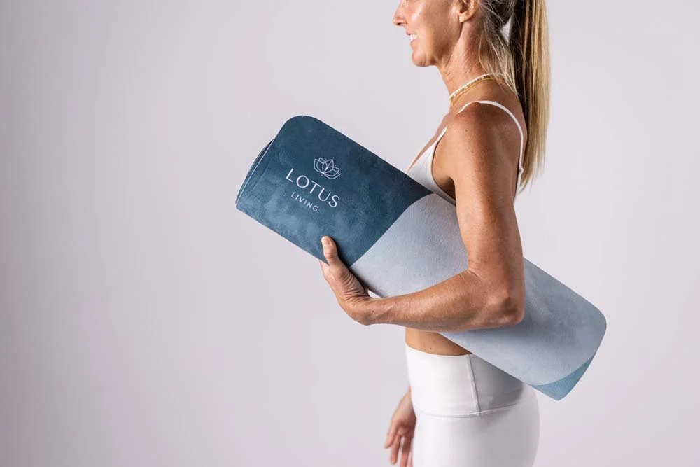 A woman in white workout attire carrying a rolled yoga mat with the logo 'Lotus Living' against a plain background.