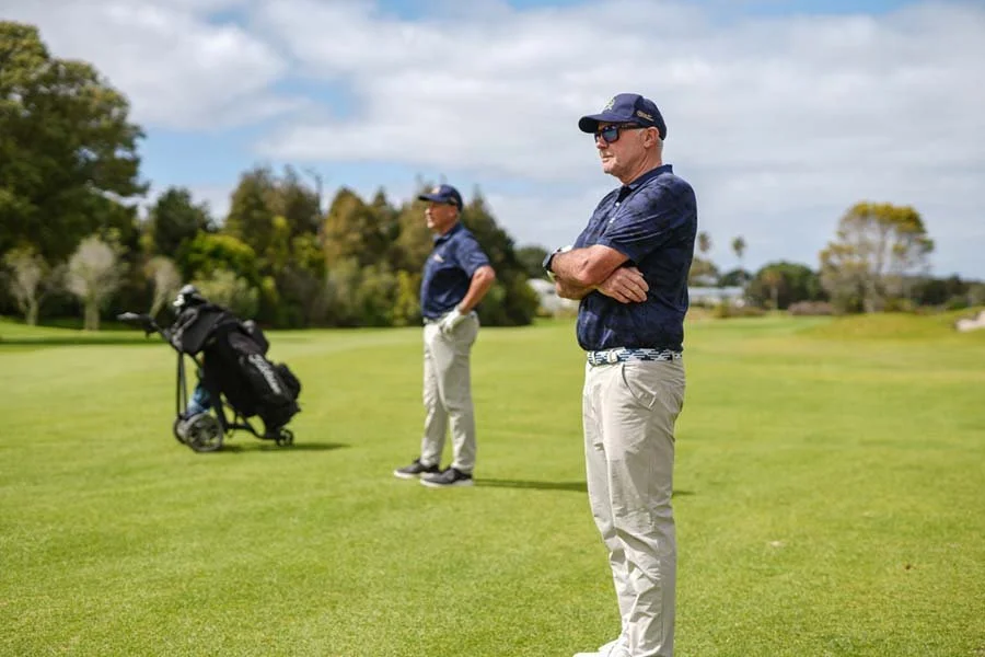Two men in golf attire stand on a golf course, one in the foreground with arms crossed, and another in the background near a golf cart with a golf bag. Trees and cloudy sky are visible in the distance.
