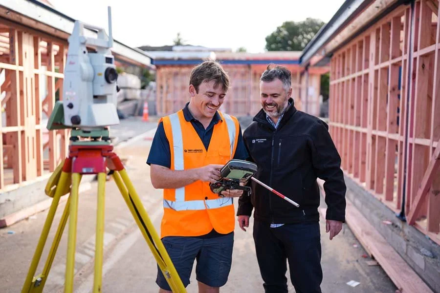 Two men at a construction site reviewing data on a tablet, with a surveying instrument on a tripod nearby, and wooden framing for a building under construction.