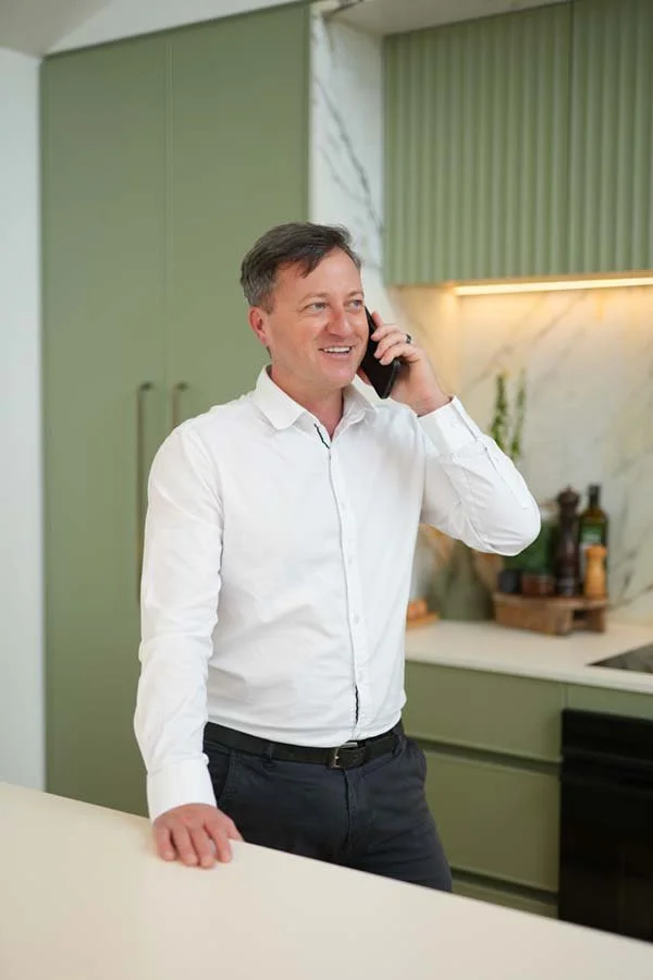 A man in a white shirt talking on a cellphone in a modern kitchen with green cabinets and a marble backsplash.