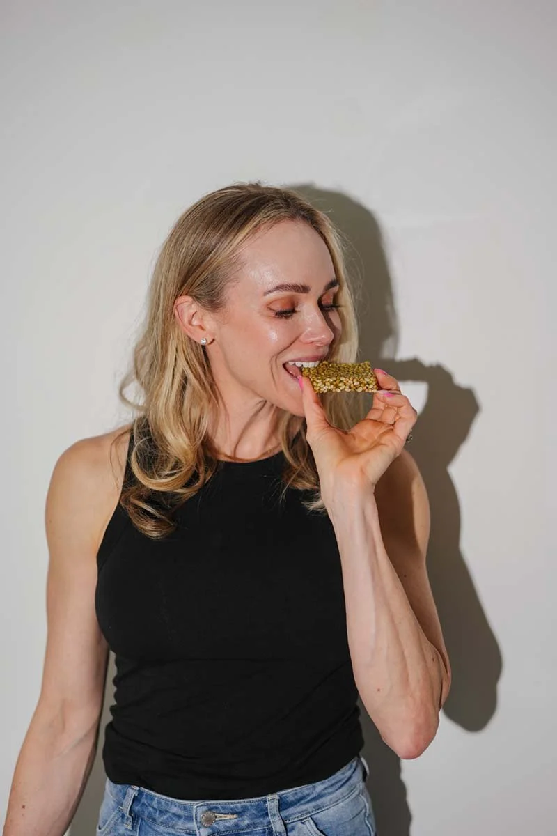 Woman with blonde hair, wearing a black sleeveless top and blue jeans, holding a sesame seed bar near her mouth as if about to eat it, standing against a plain white wall.