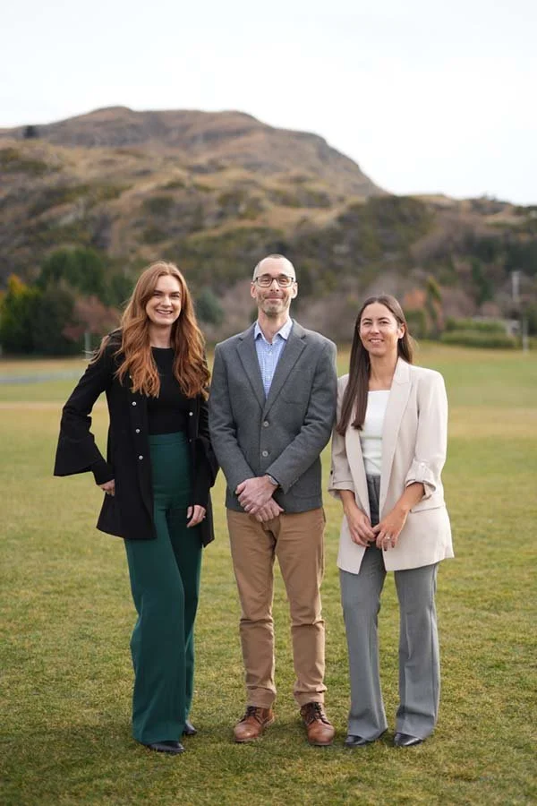 Three professionally dressed people standing on a grassy field with a mountain in the background, smiling at the camera.