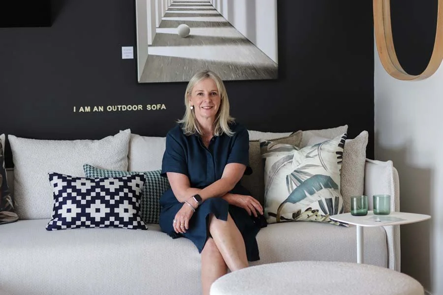 A woman sitting on a beige sofa with decorative pillows in a modern living room. Behind her is a black wall with a mirror and a wall decor that reads "I AM AN OUTDOOR SOFA." A small white side table with two green glasses is to her right.