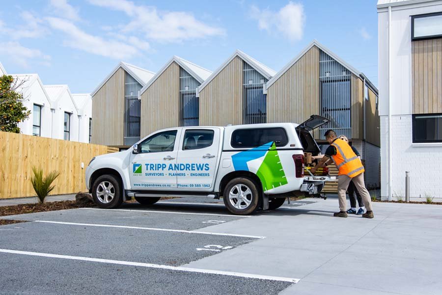 Construction worker wearing an orange safety vest loading equipment into a white survey and engineering company truck branded 'Tripp Andrews' parked in a residential area with modern houses in the background.