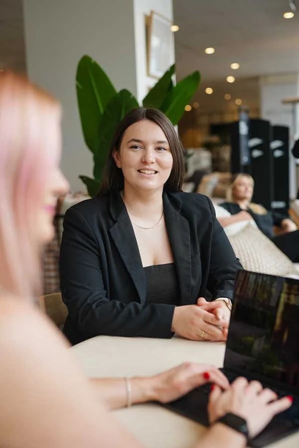 Two women sitting at a table in a modern indoor space, engaging in conversation, one in focus wearing a black blazer, the other with blonde hair working on a laptop.