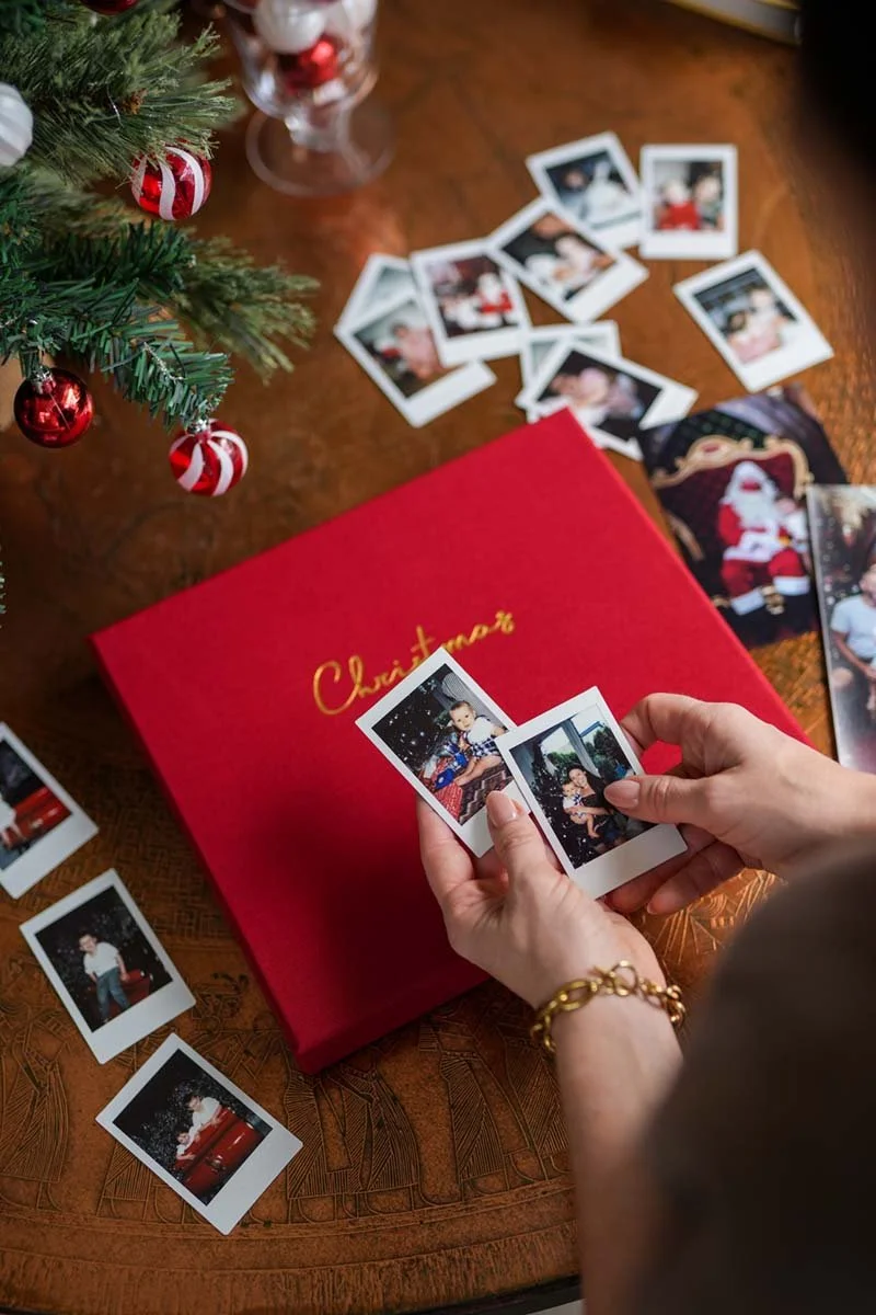 Person holding two instant photographs over a red Christmas gift box with gold writing, with scattered instant photos on a wooden table, and part of a decorated Christmas tree on the left.