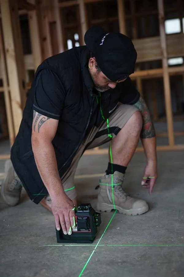 Man kneeling on the floor holding a laser level device, with green laser lines projecting onto the floor and walls of a construction site with wooden framework.