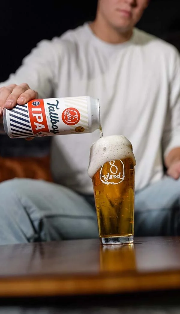 A man in a white shirt pours a can of Royal Tallboy PBR beer into a glass with foam on top.