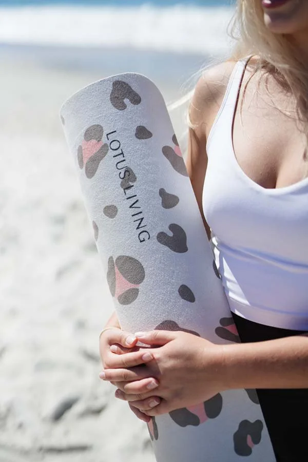A woman holding a yoga mat with a leopard print pattern and the words 'LOTS of LIVING' on it, standing on a sandy beach near the water.