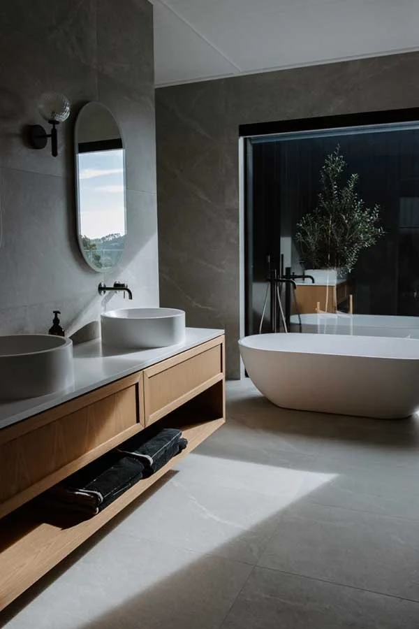 Modern bathroom with double vessel sinks, a large mirror, a freestanding bathtub by a sliding glass door, a potted plant, and wooden vanity with folded towels.