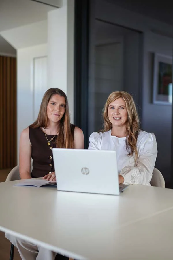 Two women sitting at a table with a laptop and a notebook, smiling, in a modern office setting.