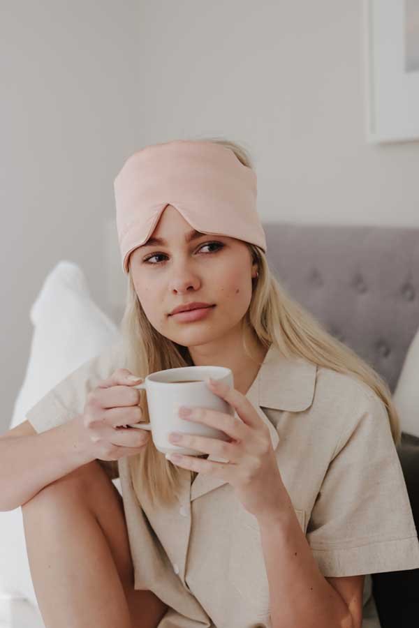 Young woman with long blonde hair wearing a pink sleep mask on her forehead, sitting on a bed holding a white mug, looking thoughtful.