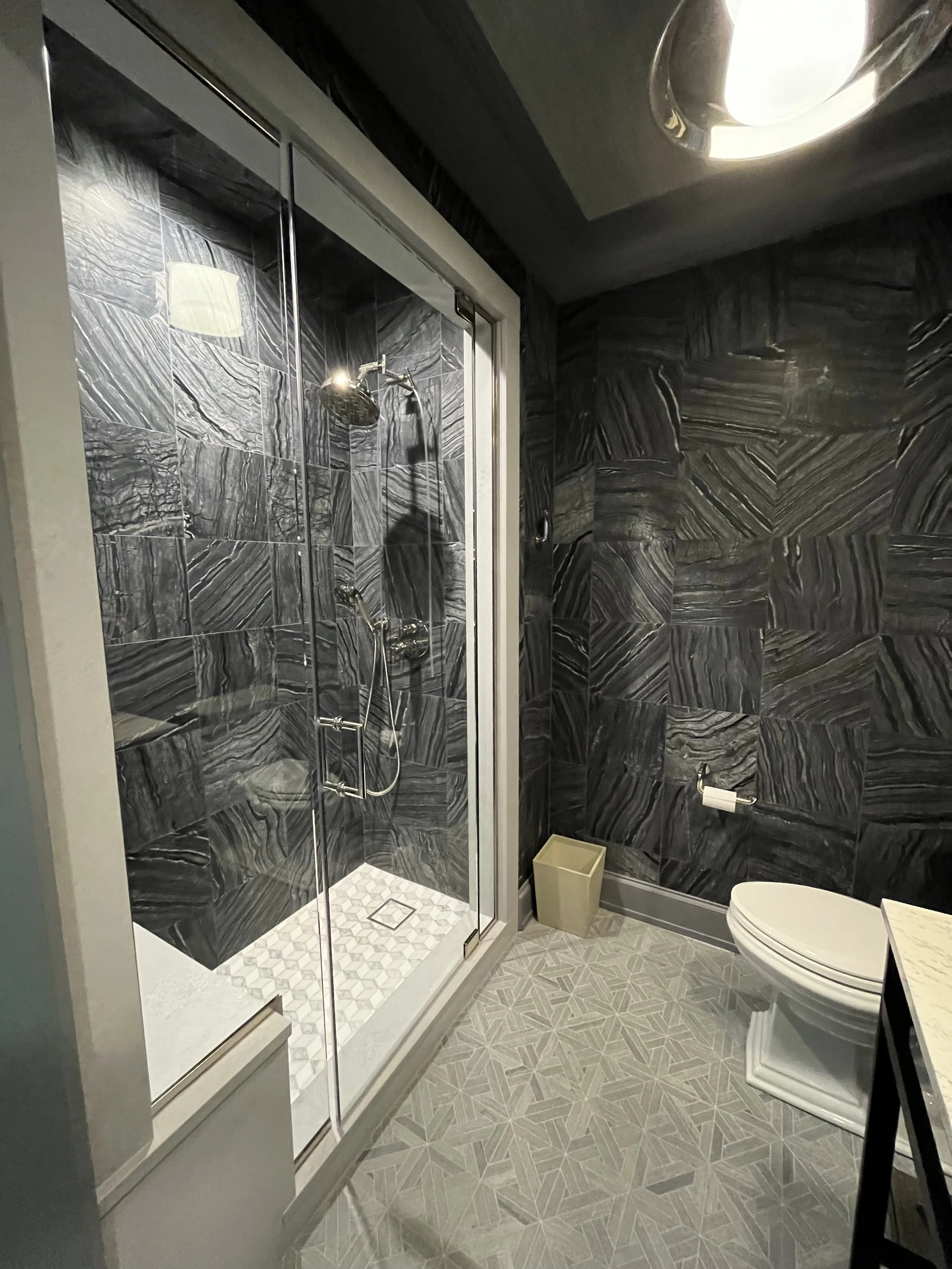 Modern bathroom with black marble walls, a glass shower enclosure, a white toilet, a small trash bin, and patterned floor tiles.