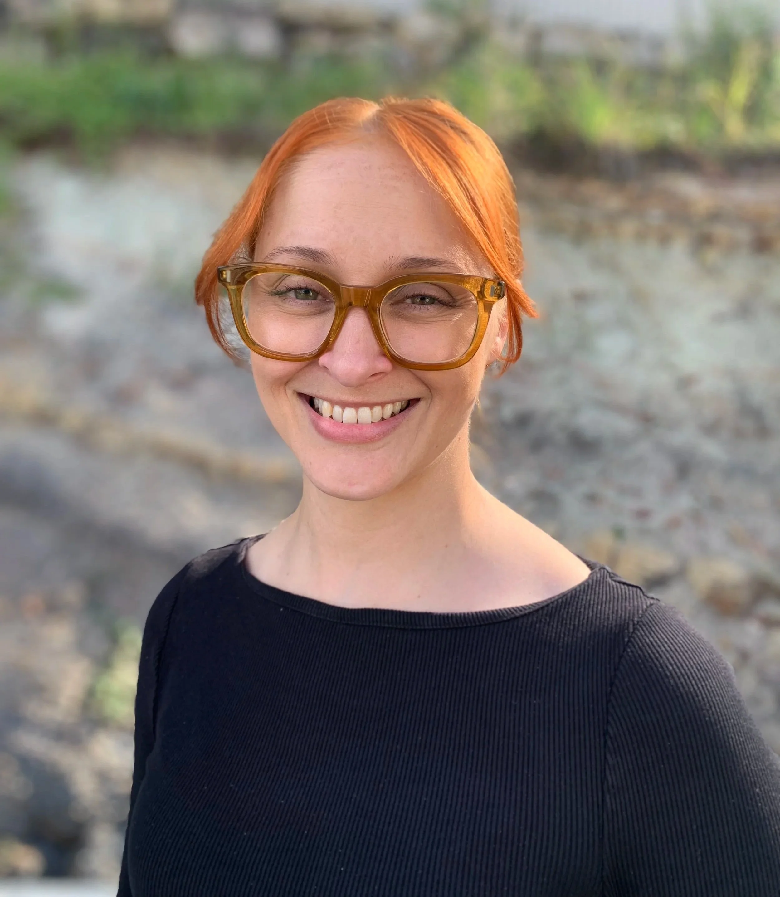 A woman with short red hair and large yellow glasses smiling outdoors with a blurred natural background Lactology Sunshine Coast.