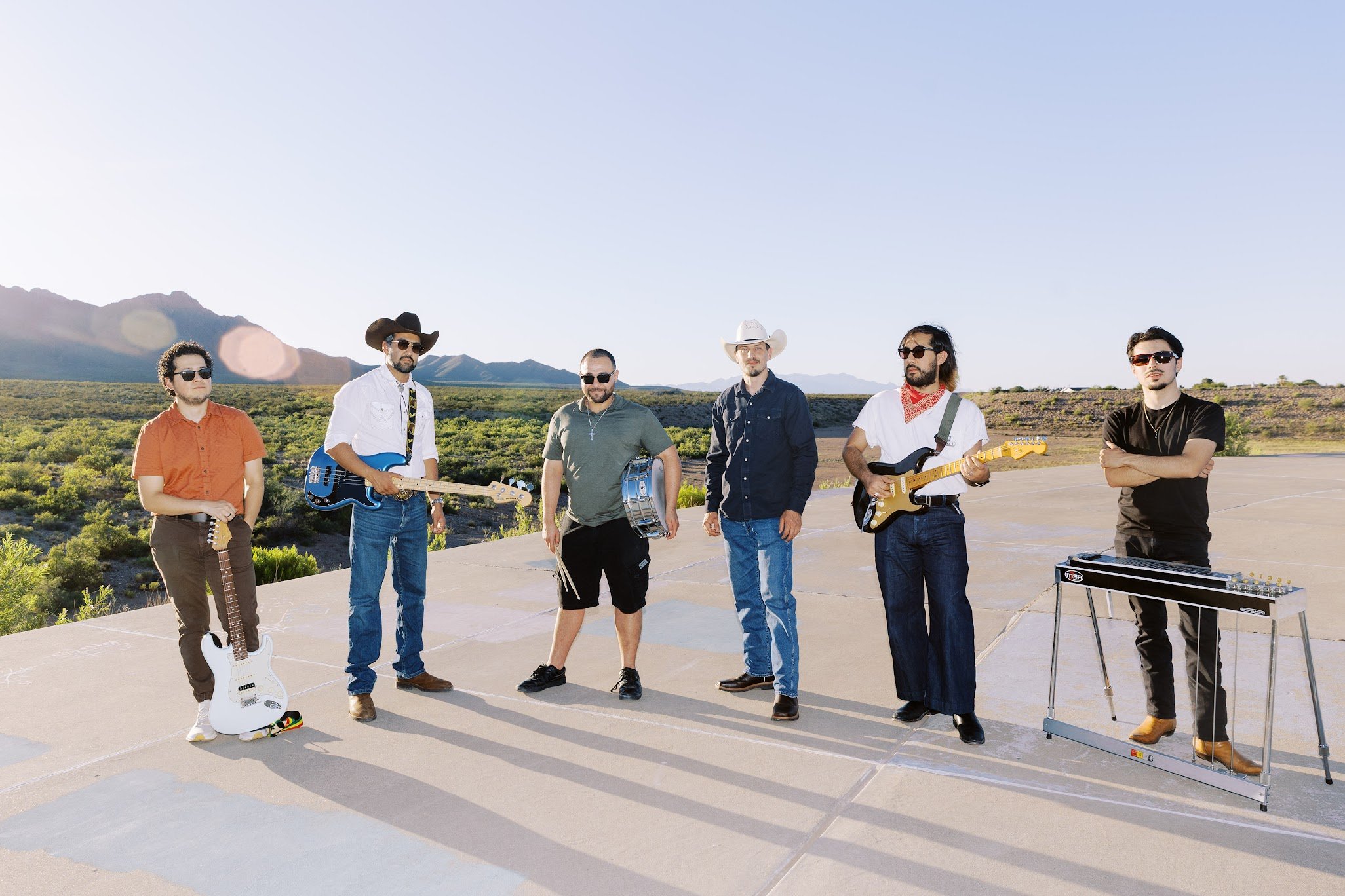 Group of six men standing outdoors on a concrete surface, holding musical instruments, with mountains and open land in the background under a clear blue sky.