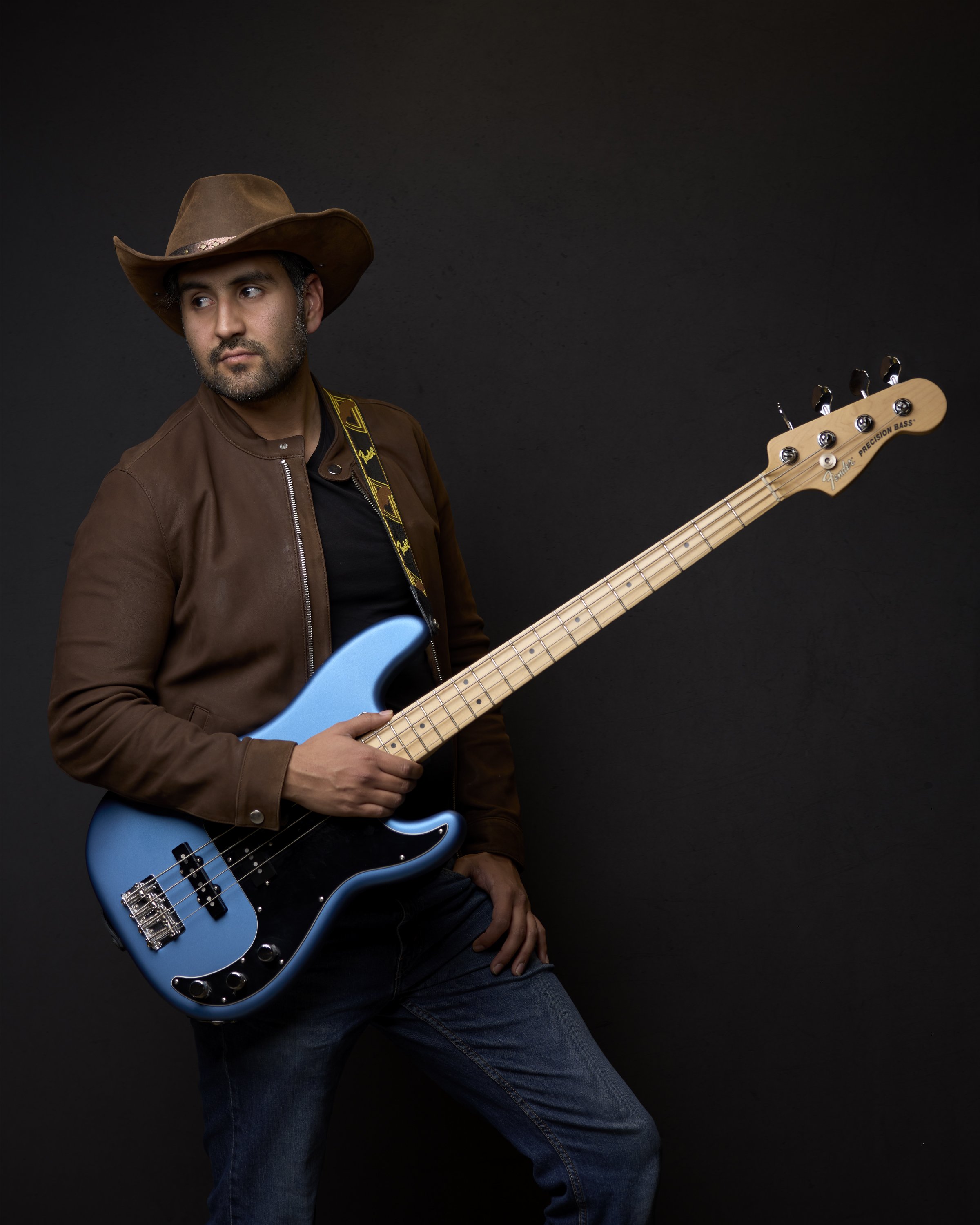 A man wearing a brown leather jacket and cowboy hat holding a blue electric bass guitar against a black background.