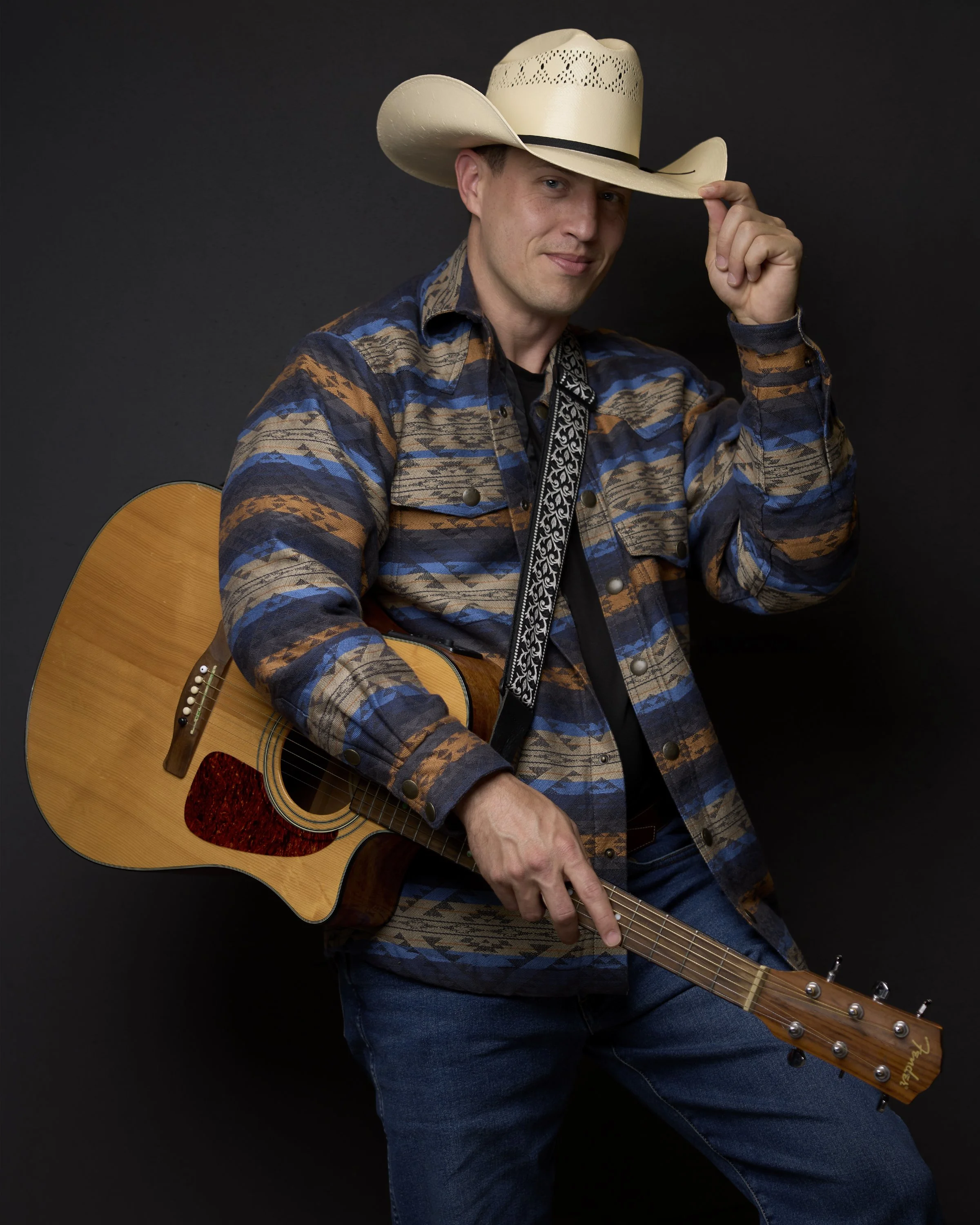 A man wearing a cowboy hat, patterned shirt, dark jeans, and holding an acoustic guitar against a black background.