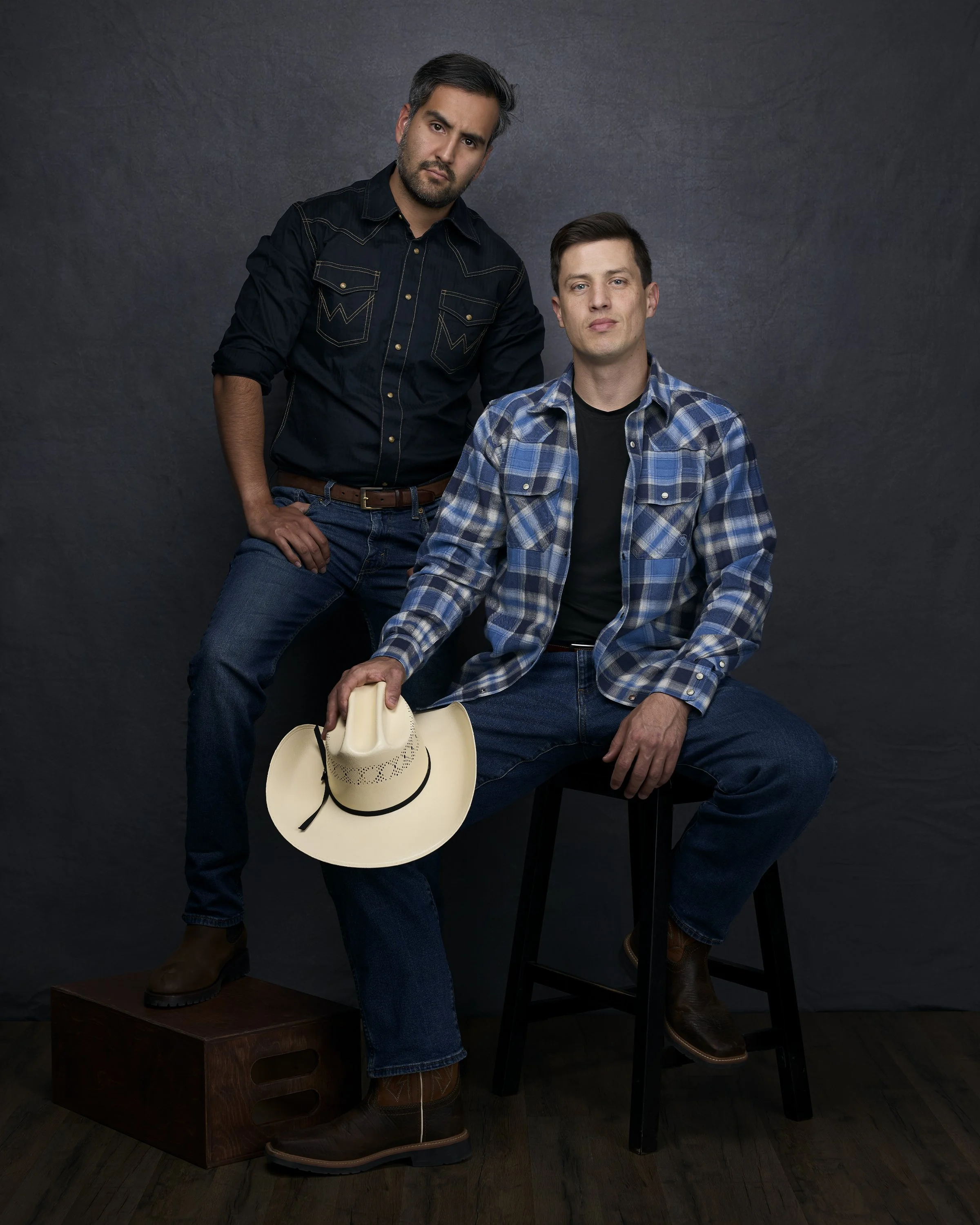 Two men dressed in jeans and plaid shirts, one seated on a stool holding a cowboy hat, the other standing with one hand on his hip, against a dark background.