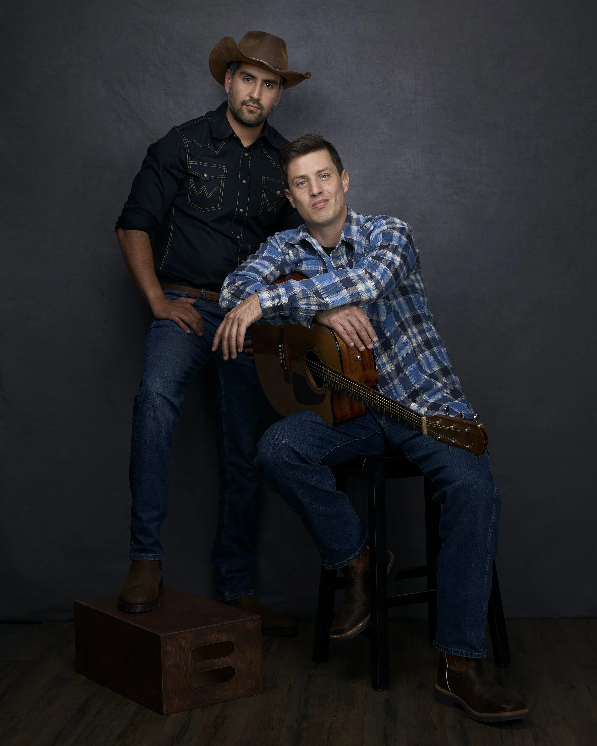 Two men with one playing an acoustic guitar and the other standing behind him. They are wearing casual western-style clothing, including plaid shirts and jeans, against a dark background.