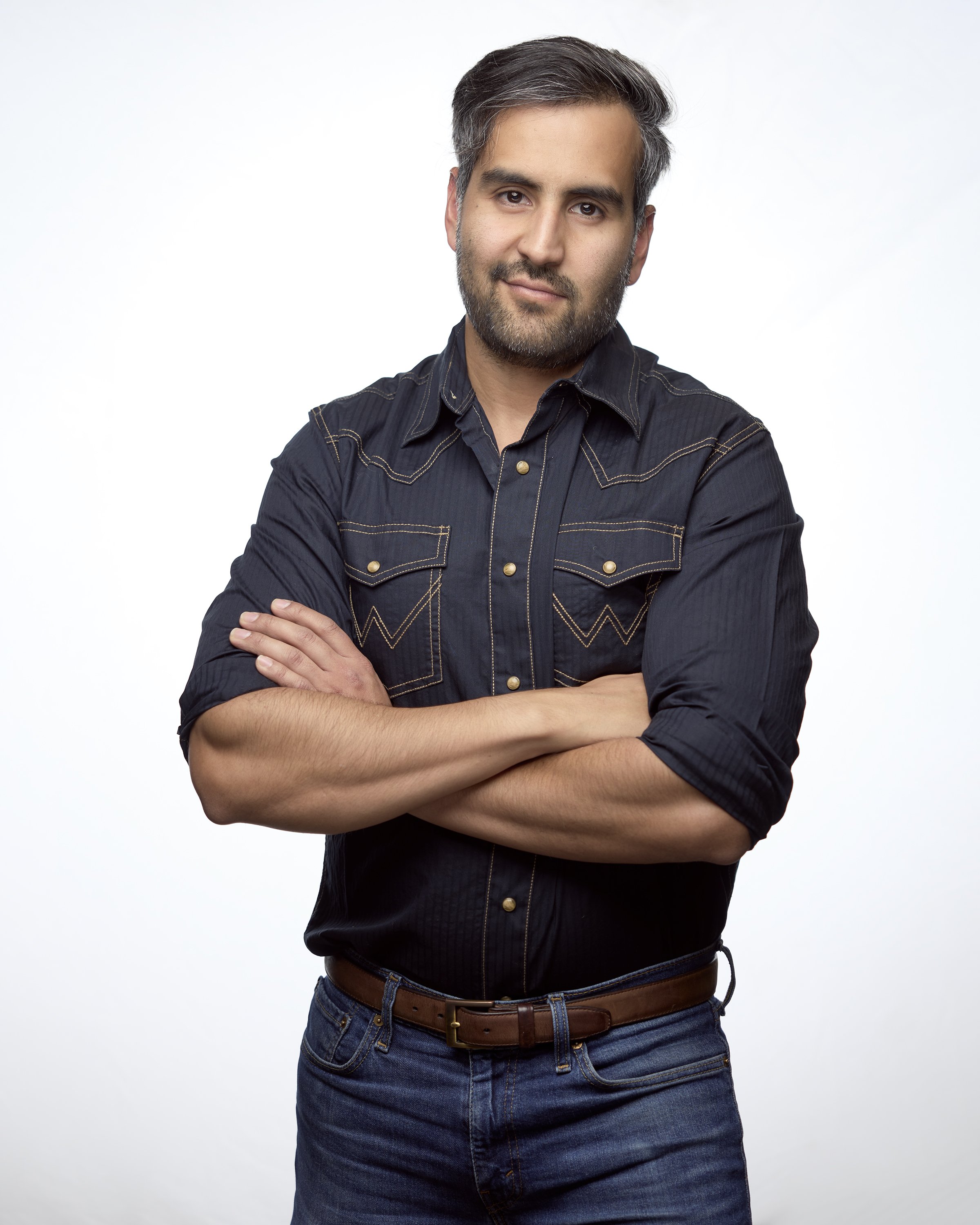 A man with gray hair and a beard, wearing a black button-up shirt with yellow stitching, standing with arms crossed against a white background.