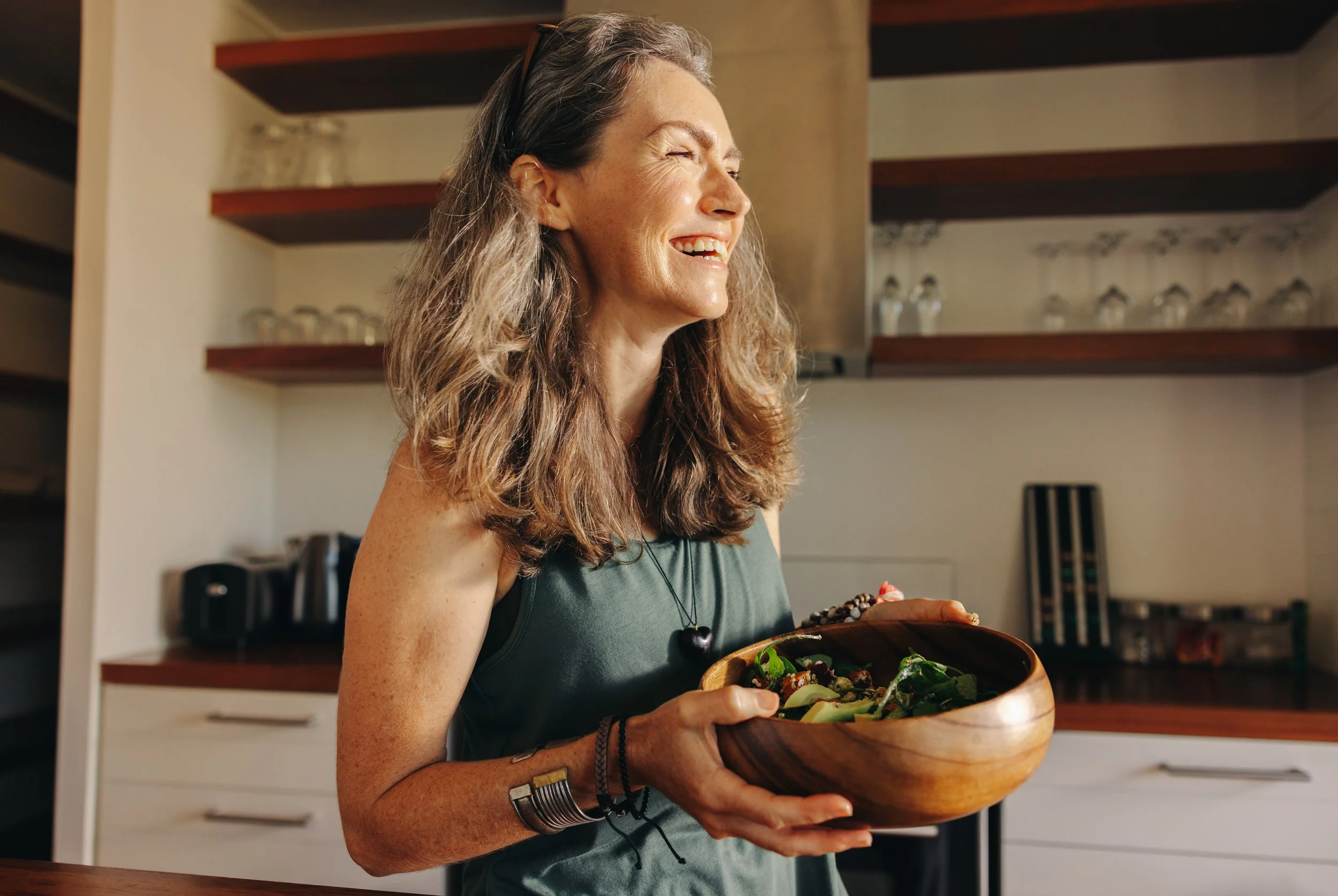 A woman with gray hair and a sleeveless top smiling and holding a wooden bowl of salad in a kitchen.