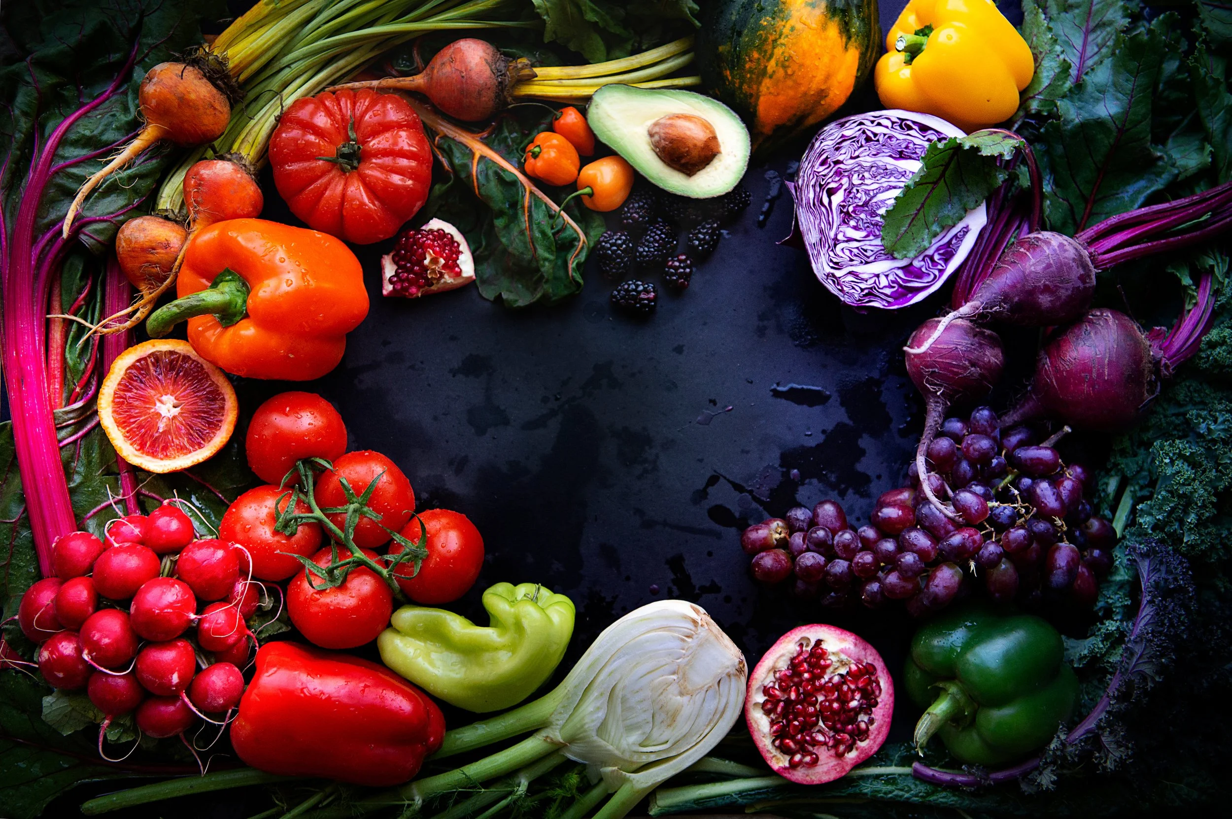 Colorful assortment of fresh vegetables and fruits arranged in a circular pattern on a black surface, including tomatoes, bell peppers, grapes, pumpkin, avocado, radicchio, beets, strawberries, blood orange, and leafy greens.