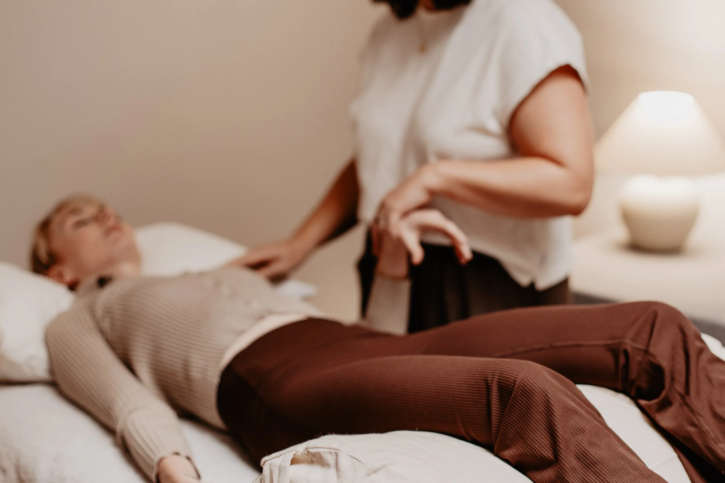 A woman lying on a bed with a young girl. The woman is reaching out and holding the girl's hand. The girl is resting on the bed, looking up. The scene appears to be indoors, with a lit lamp and some decorative vases in the background.