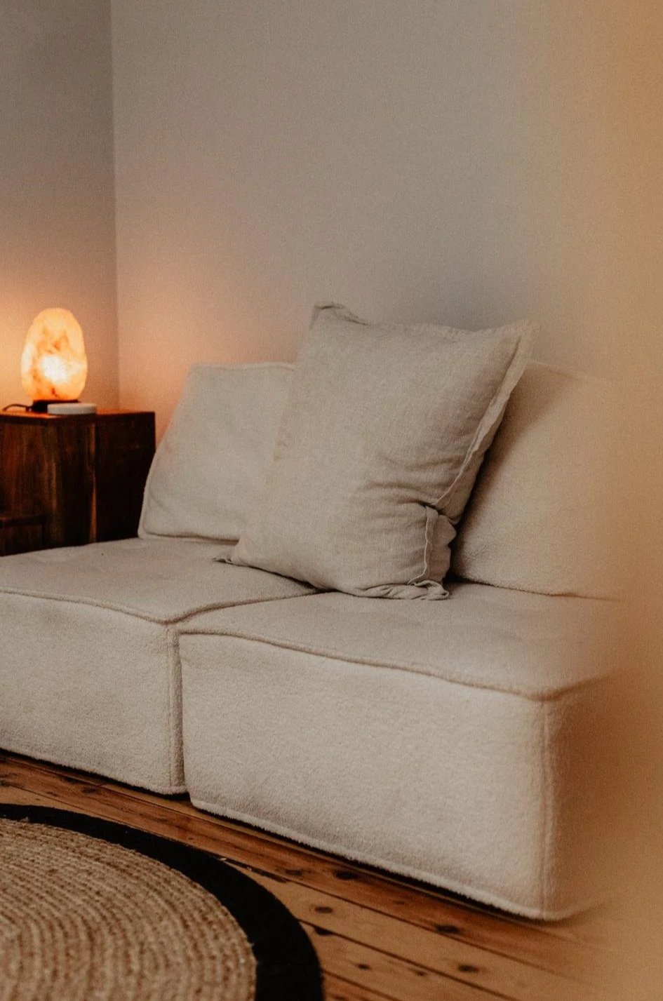 A beige sofa with two large cushions in a cozy living room corner, next to a wooden side table with a Himalayan salt lamp emitting warm light, and a woven rug on a wooden floor.