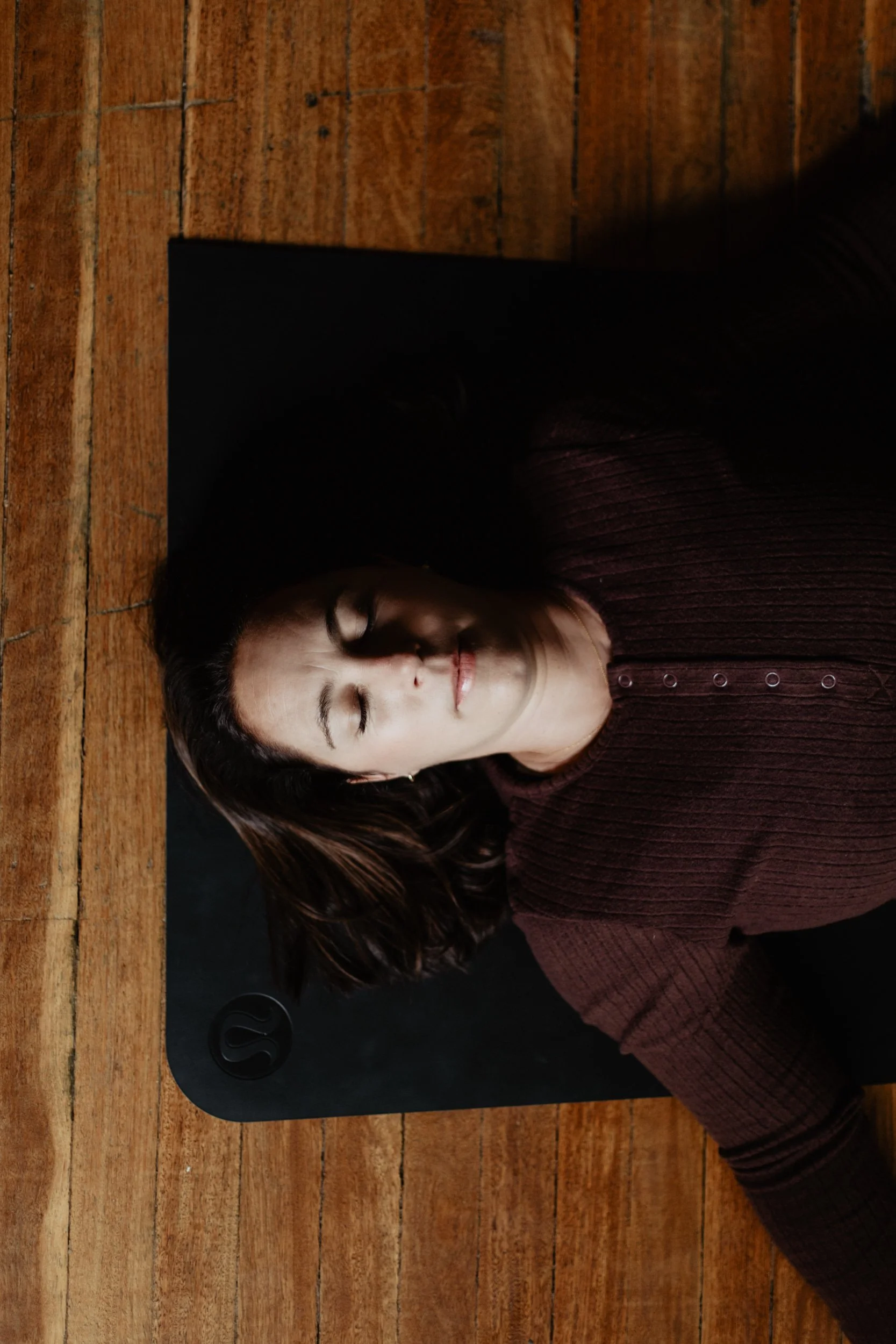 Woman with long dark hair lying on a wooden floor, resting her head on a black laptop with a logo on it, wearing a dark red sweater with buttons.