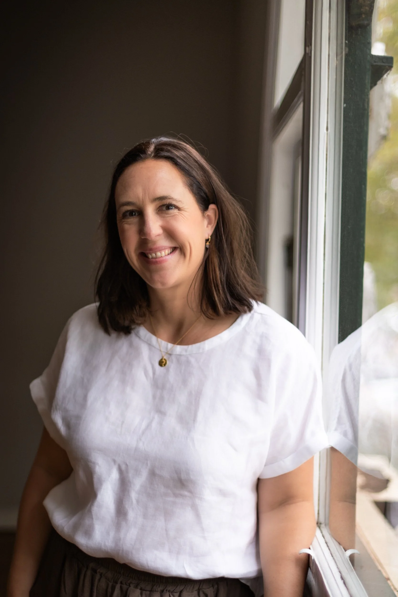 A woman with shoulder-length brown hair, smiling, standing by a window in a room, wearing a white blouse and gold jewelry.
