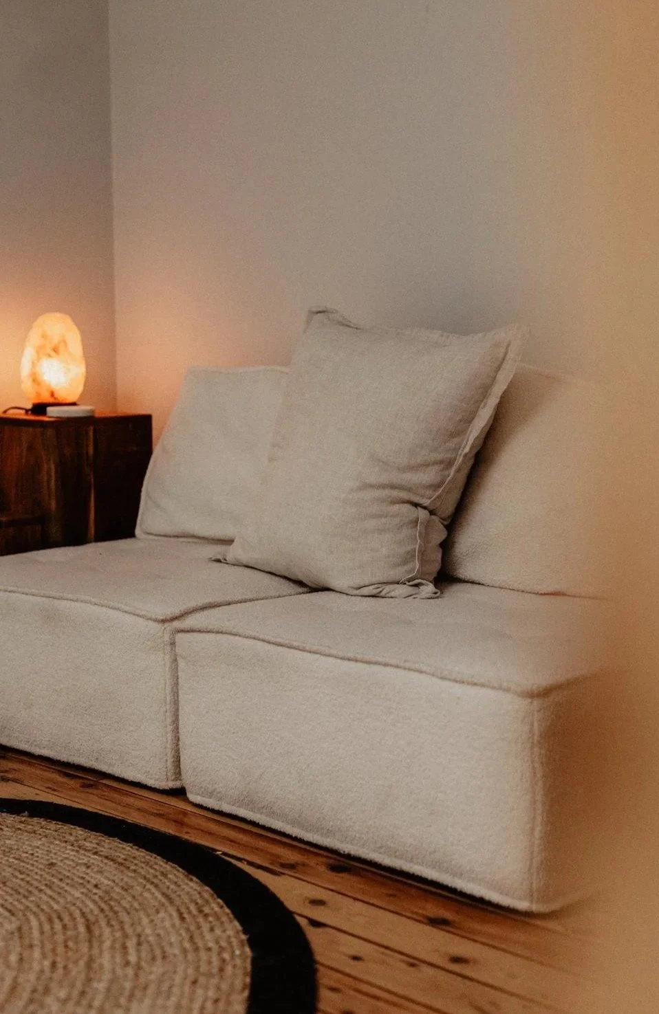 A beige sofa with two matching pillows in a cozy living room corner, next to a wooden side table with a salt lamp and a small object, on a wooden floor with a woven circular rug.