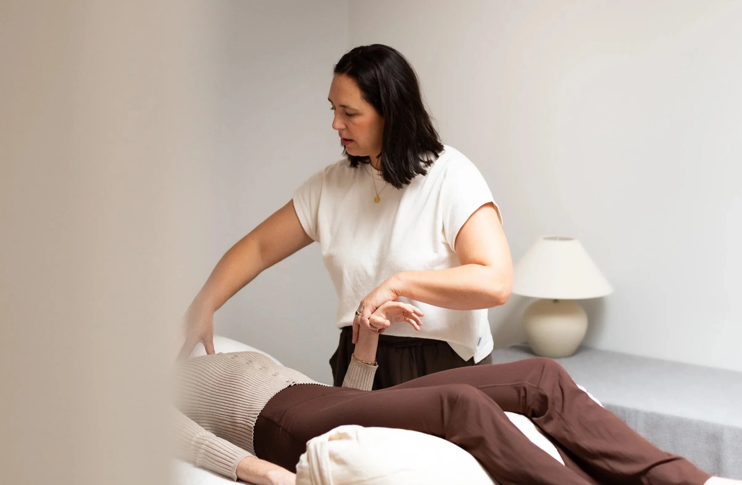 A woman with dark hair and wearing a white t-shirt is performing a massage or physical therapy on a patient lying on a massage table or bed, wearing beige sweater and brown pants, in a minimally decorated room with a lamp on a side table.