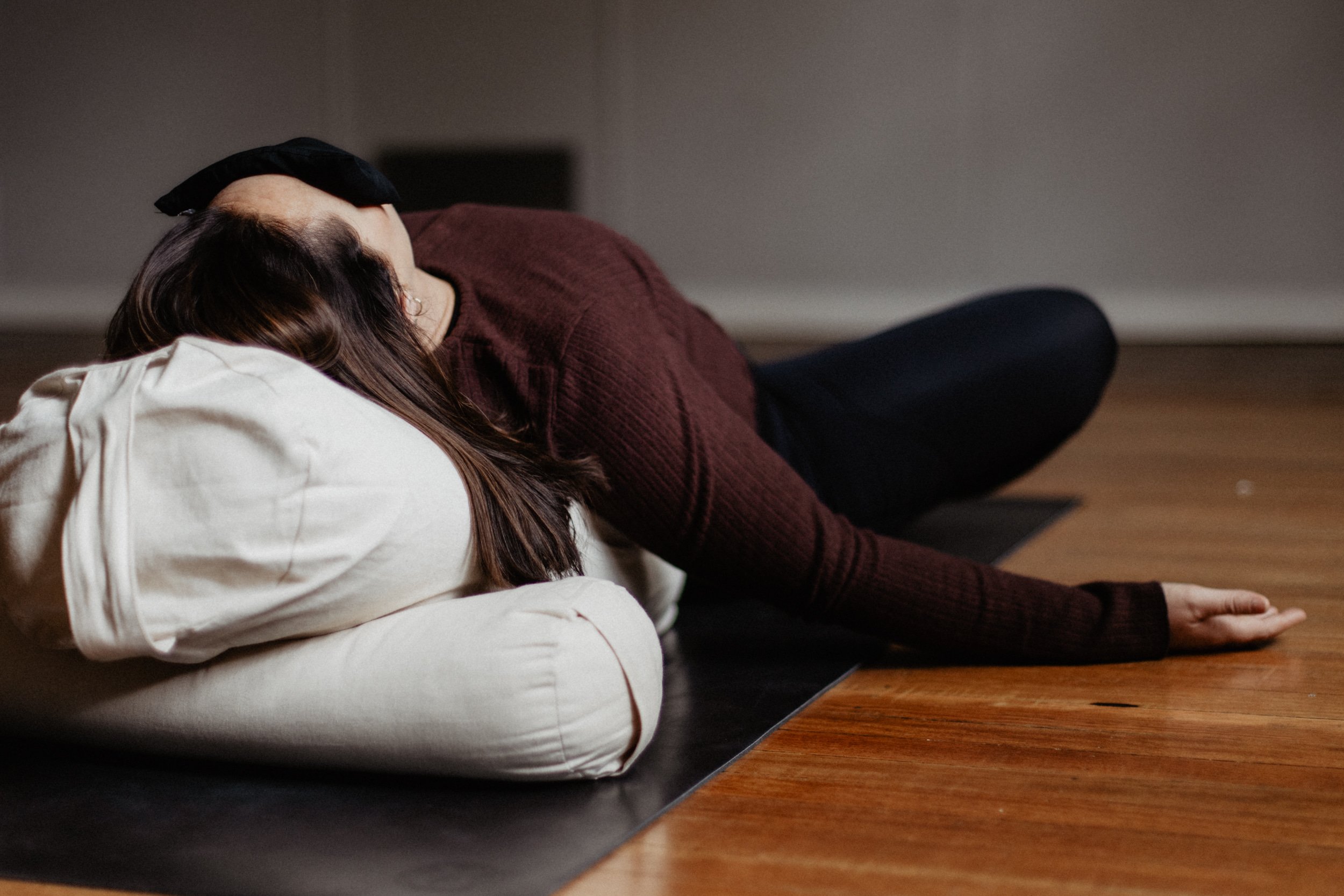Person practicing yoga or meditation on a yoga mat, lying on their side with eyes closed and head resting on a pillow, in a room with a wooden floor.