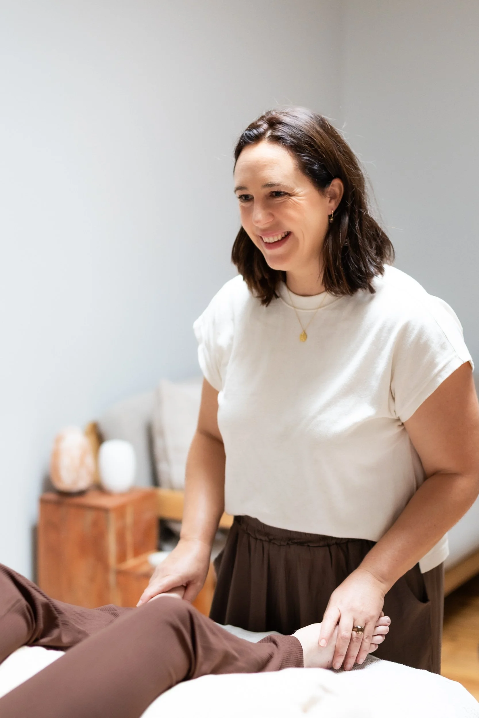 A woman smiling while holding a person's hand in a room with neutral walls and wooden furniture.