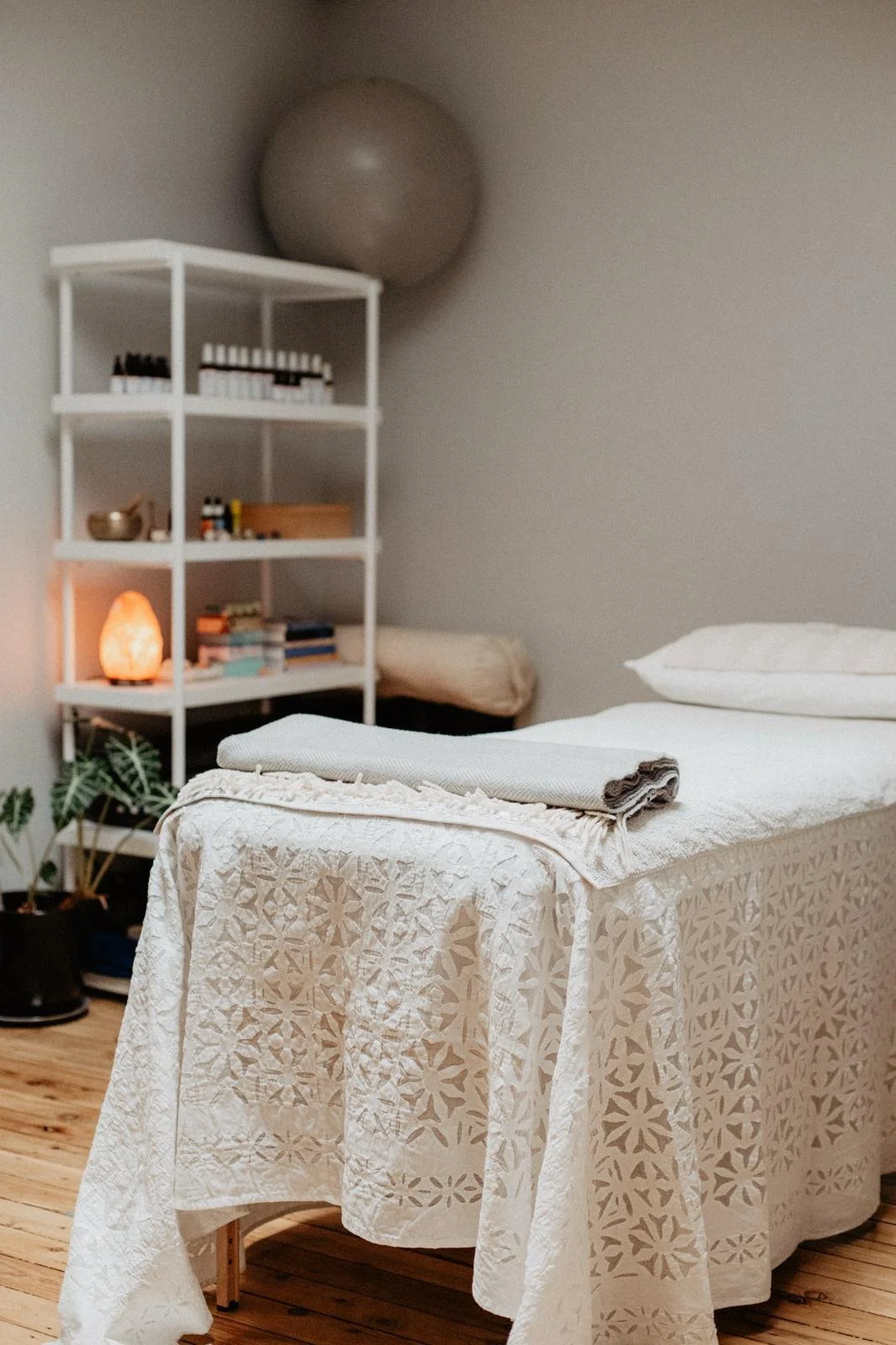 A cozy wellness or massage room with a bed covered by a decorative white bedspread, a folded towel on top, a white pillow, a salt lamp, and a bookshelf in the background filled with books, essential oils, and a salt lamp, a large exercise ball on the wall, and a potted plant on the floor.
