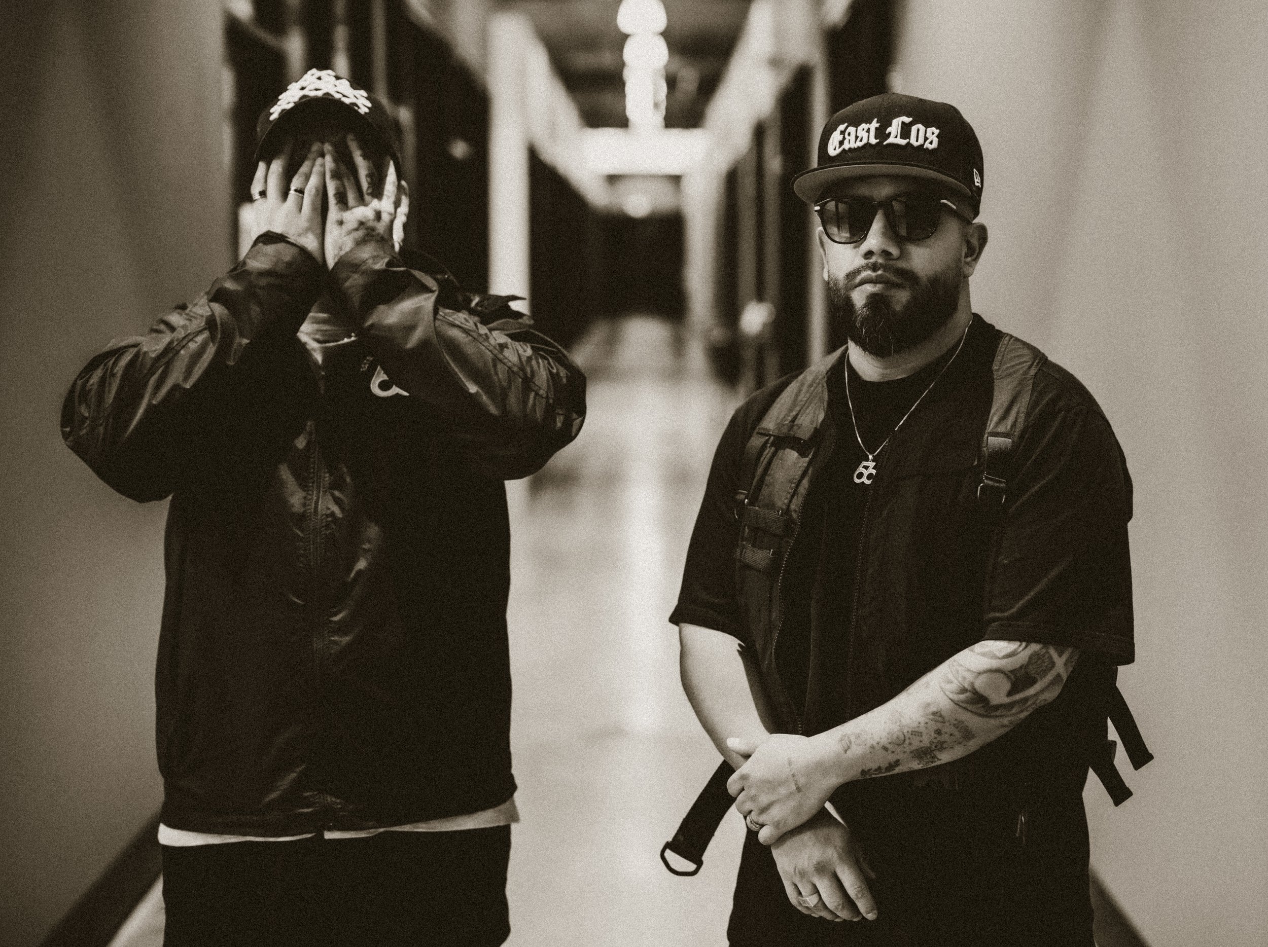 Two men standing in a corridor, one covering his face with his hands and the other looking at the camera wearing sunglasses and a cap that says 'East Los'. The man on the right has tattoos on his arm, a beard, and a backpack.
