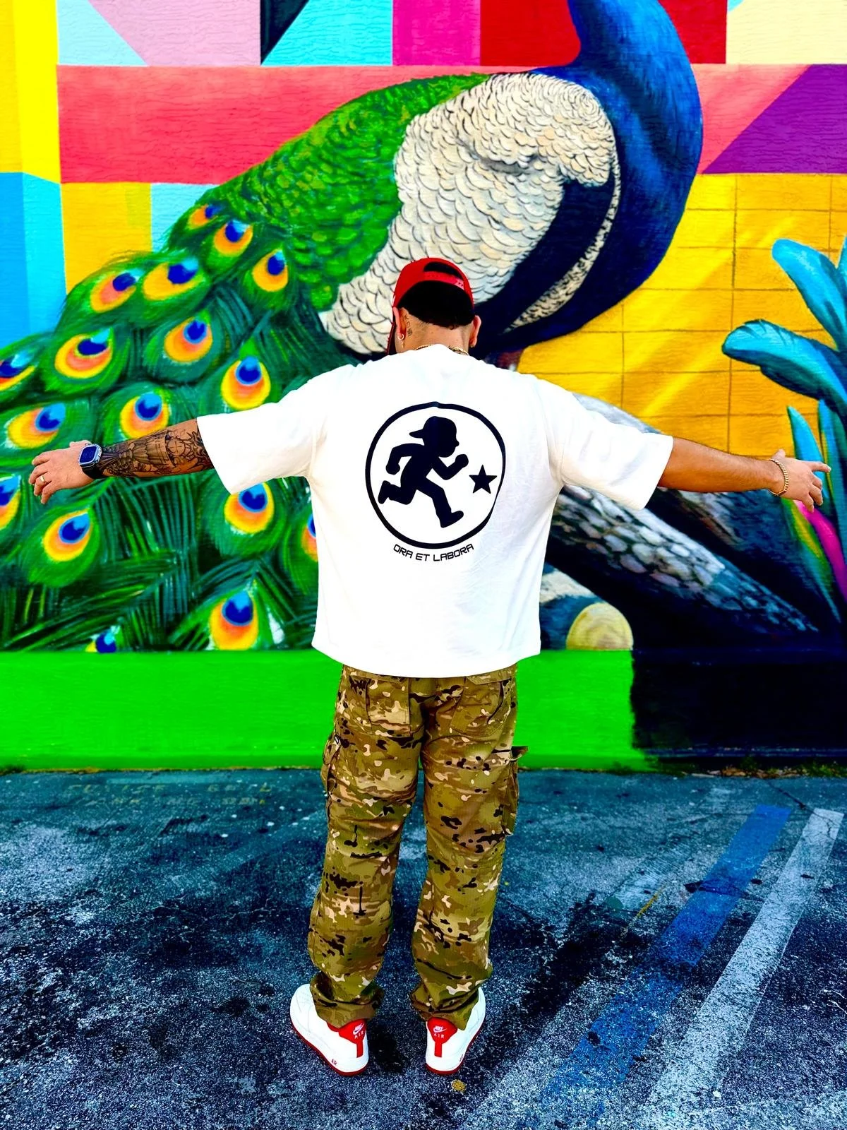 A person with tattoos on their left arm, wearing a red cap backwards, white shirt with a black logo of a running figure and Latin phrase, camouflage pants, and white sneakers with red accents, stands with arms outstretched in front of a colorful mural of a peacock with vibrant feathers in green, blue, and yellow, on a city street.
