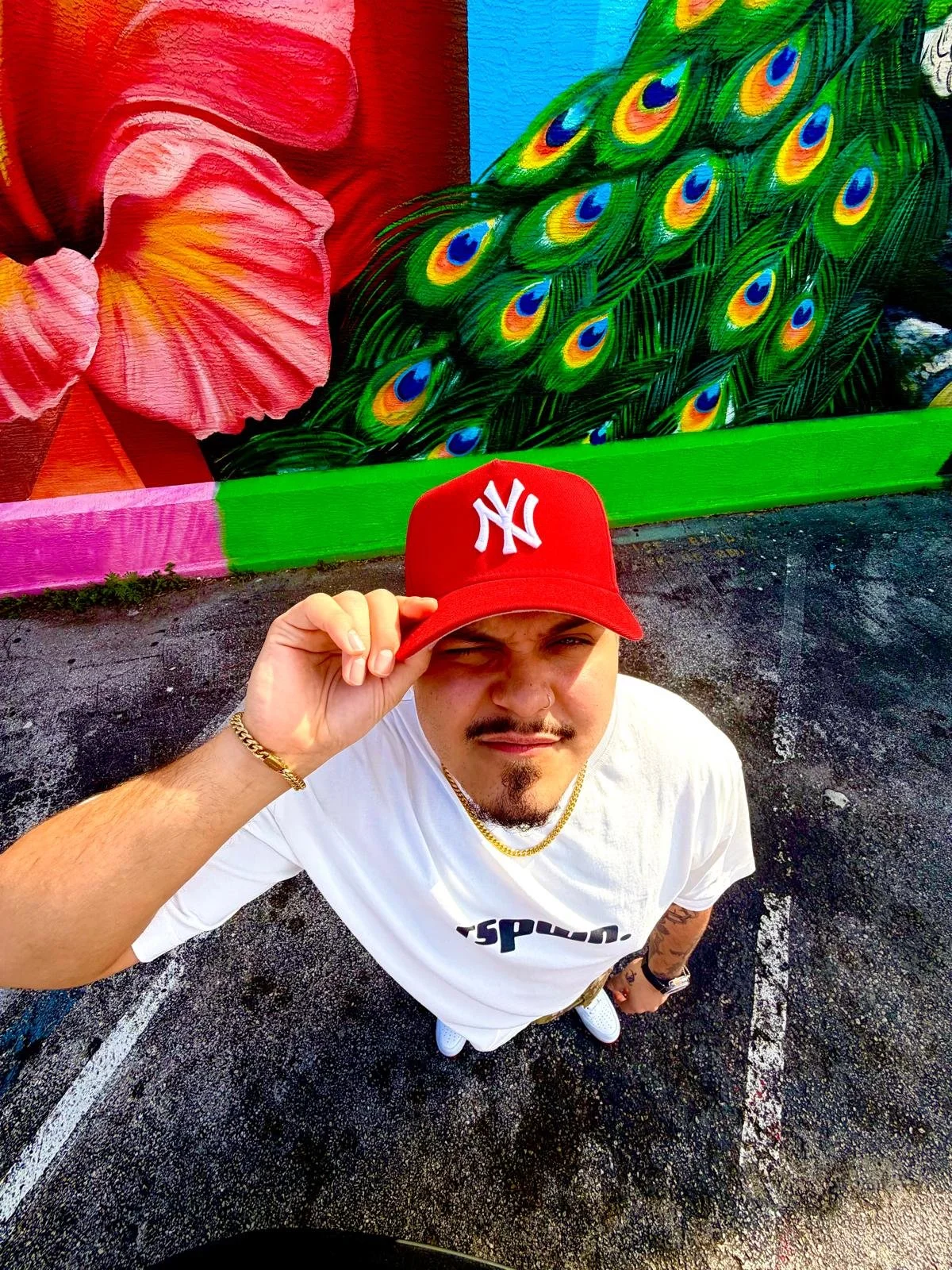 Young man wearing a red New York Yankees cap, white t-shirt, gold chain, and bracelet, taking a selfie in front of a colorful mural featuring large pink flowers and a peacock with vibrant feathers.