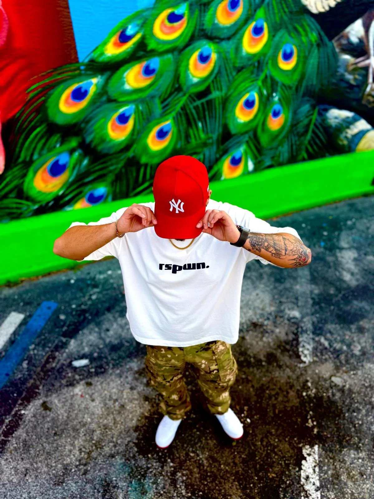 A person standing in a parking lot, wearing a red baseball cap with the Yankees logo, a white t-shirt with black text, camouflage cargo pants, and white shoes. They are adjusting their cap and have tattoos on one arm. Behind them, there is a colorful mural of peacock feathers on a wall.
