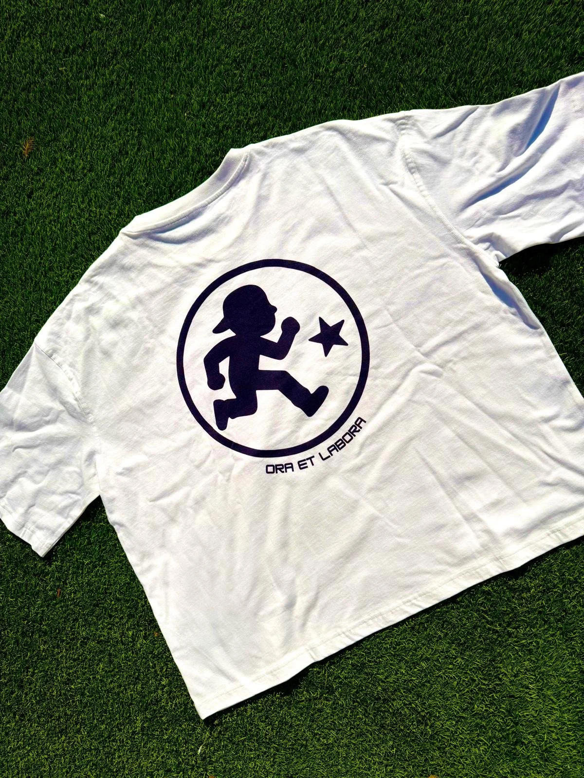 White T-shirt with a black graphic of a running child wearing a hat, inside a circle, with a star to the right of the child, and the Latin phrase "ORA ET LABORA" written below.