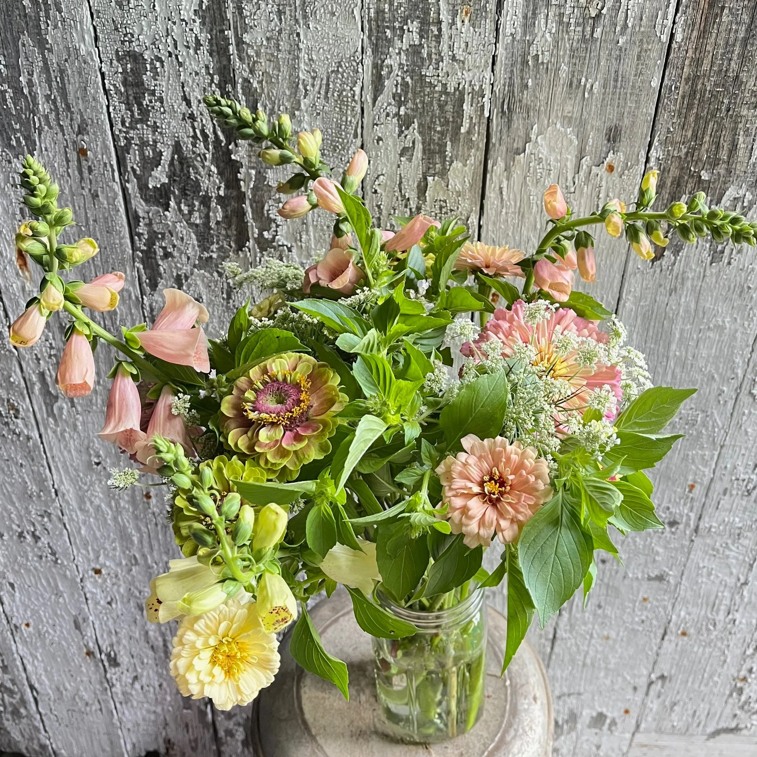 A bouquet of assorted pastel-colored flowers in a glass jar on a small round table against a weathered wooden background.