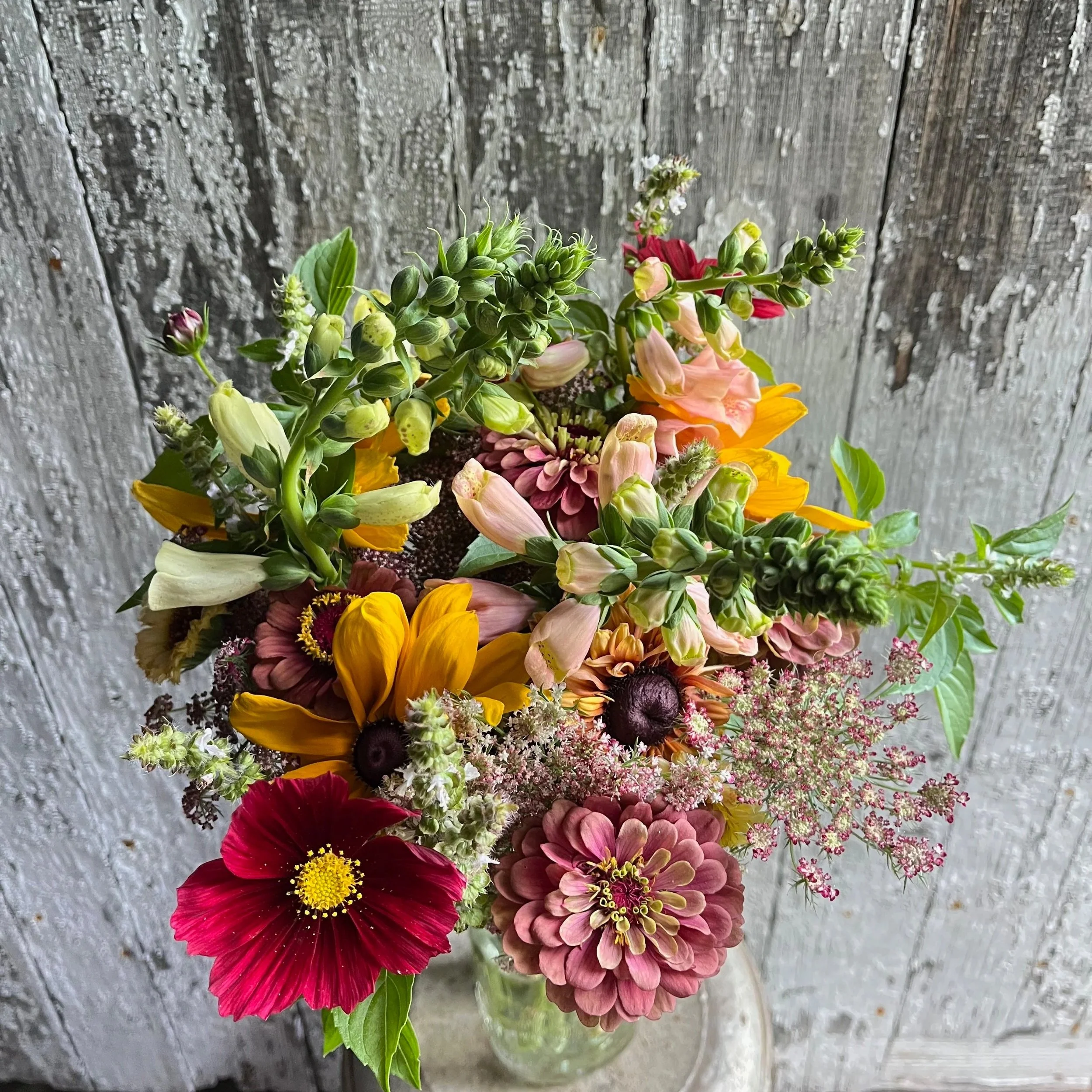 Colorful bouquet of flowers including sunflowers, dahlias, cosmos, and other mixed flowers in a glass vase against a rustic wooden backdrop.