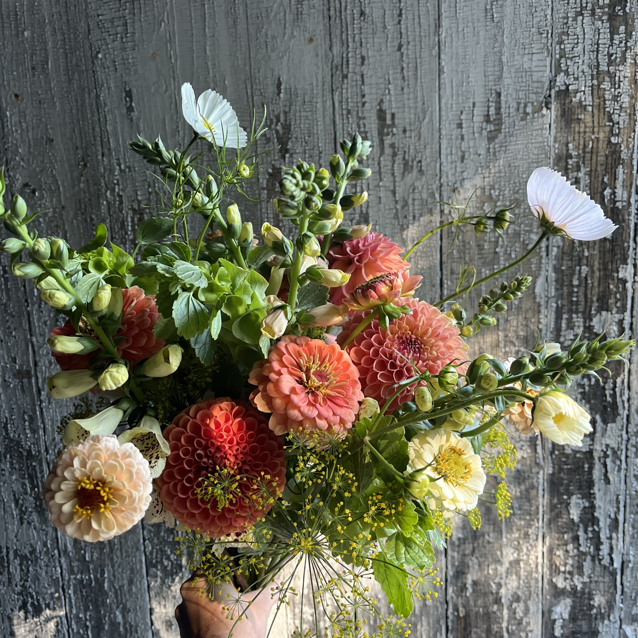 A colorful bouquet of flowers including dahlias, sweet peas, dill, and other blooms in a hand against a weathered wooden background.