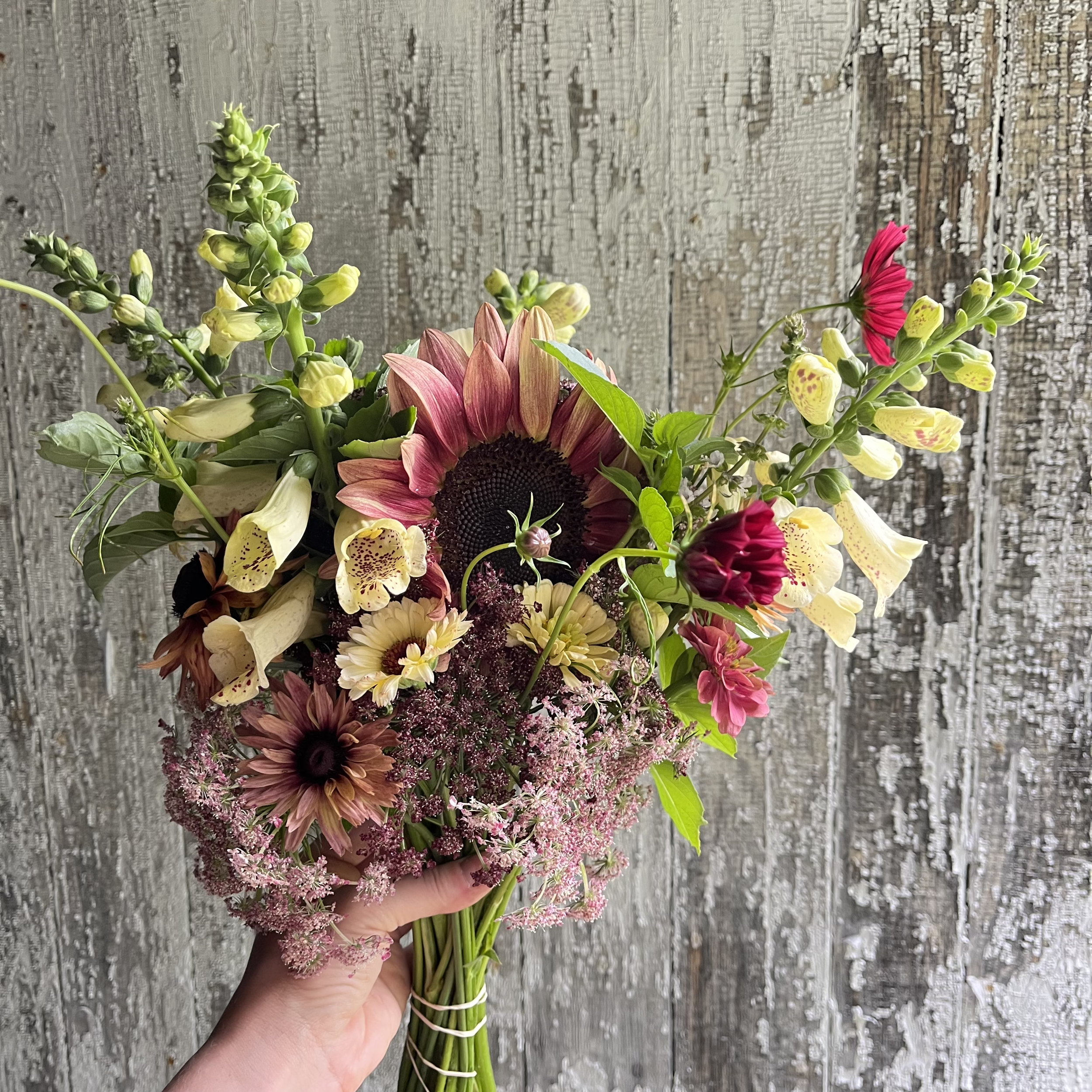 Hand holding a bouquet of mixed flowers including sunflowers, daisies, snapdragons, and others against a weathered wooden background.