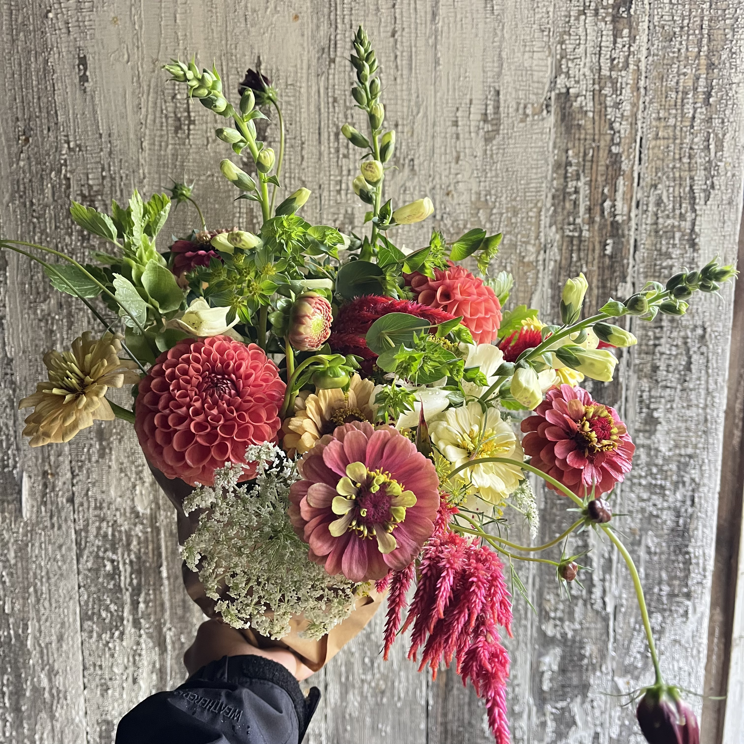 A colorful bouquet of various flowers including dahlias, snapdragons, zinnias, and other blooms, held in a person’s hand against a weathered wooden background.