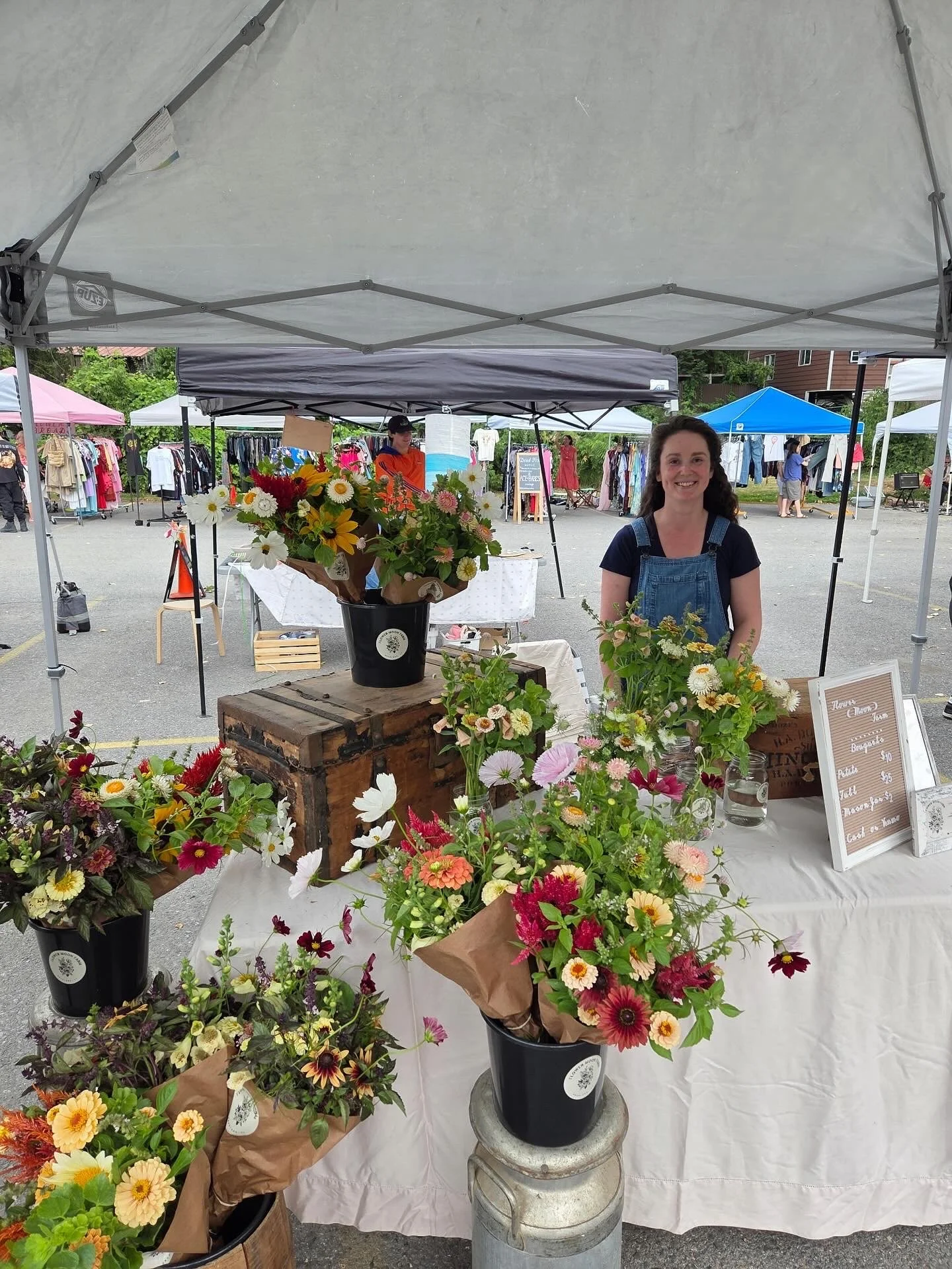 A smiling woman standing behind a table with various colorful flower arrangements at an outdoor market stall, with other market tents and shoppers visible in the background.