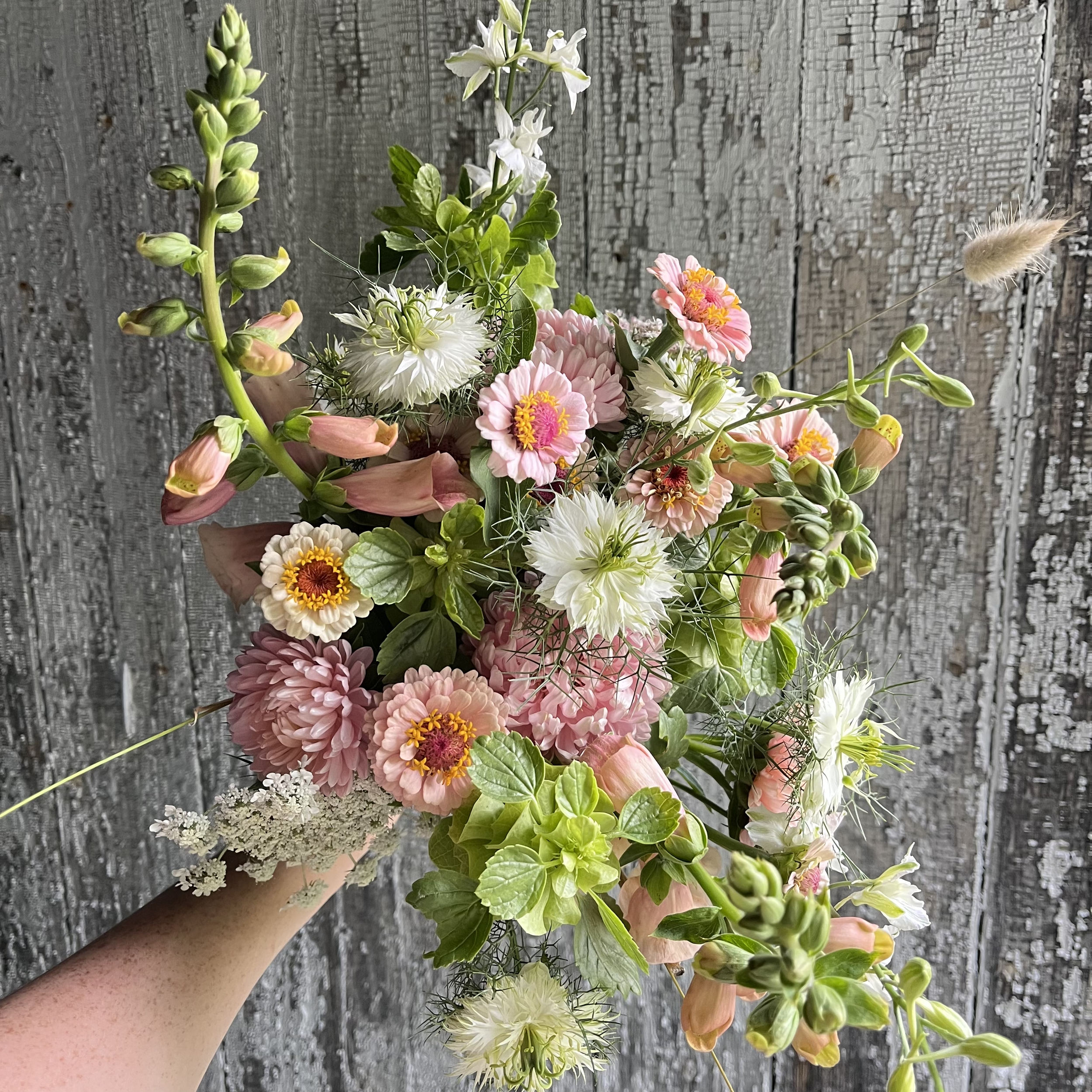 A hand holding a colorful arrangement of various flowers and greenery against a weathered wooden background.