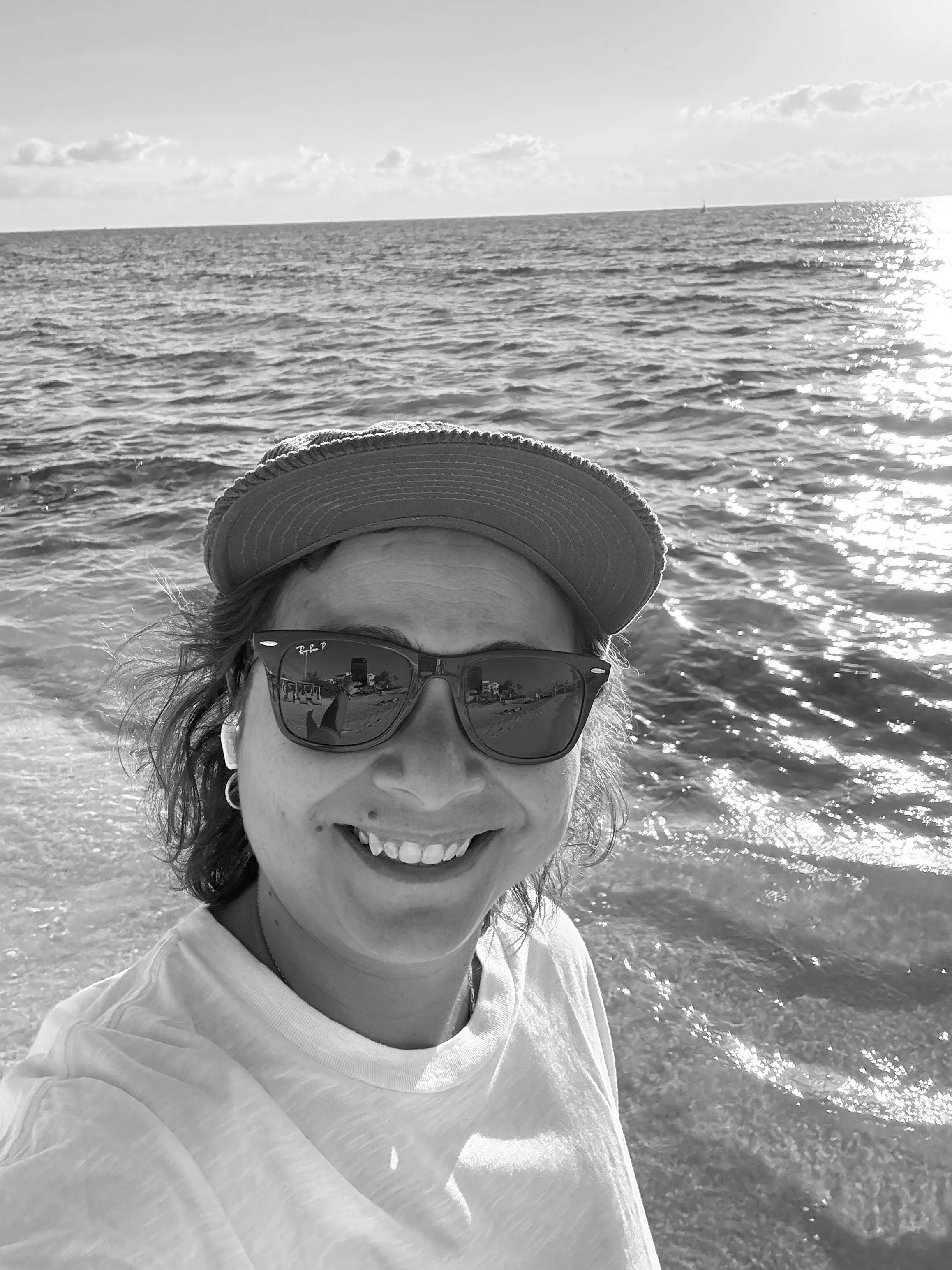 A woman taking a selfie at the beach during sunset, wearing sunglasses, a hat, and a white t-shirt, with the ocean and sky in the background.