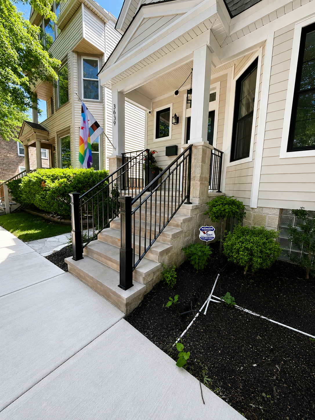 Exterior view of a house with a front porch, steps with black metal railing, and a garden with bushes and plants. A rainbow pride flag is visible near the front entrance.